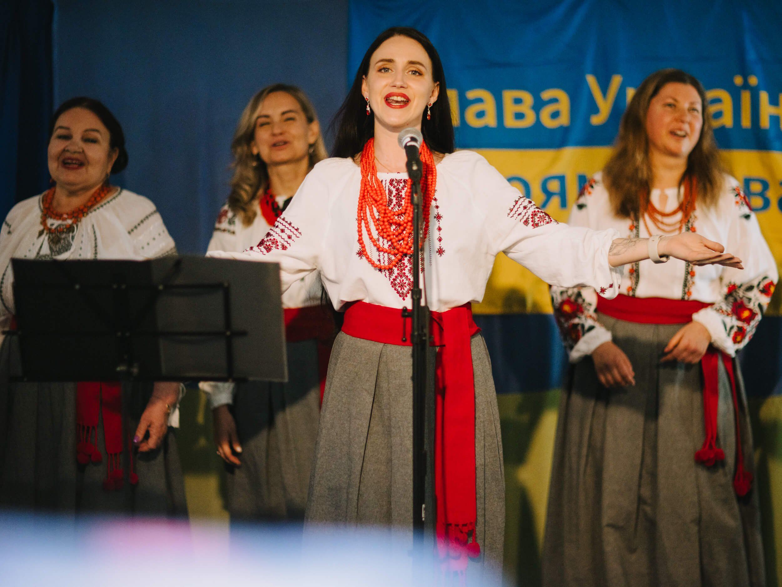 Four women in traditional Ukrainian embroidered clothing perform on stage, the center woman singing at a microphone with red lipstick and a bright smile