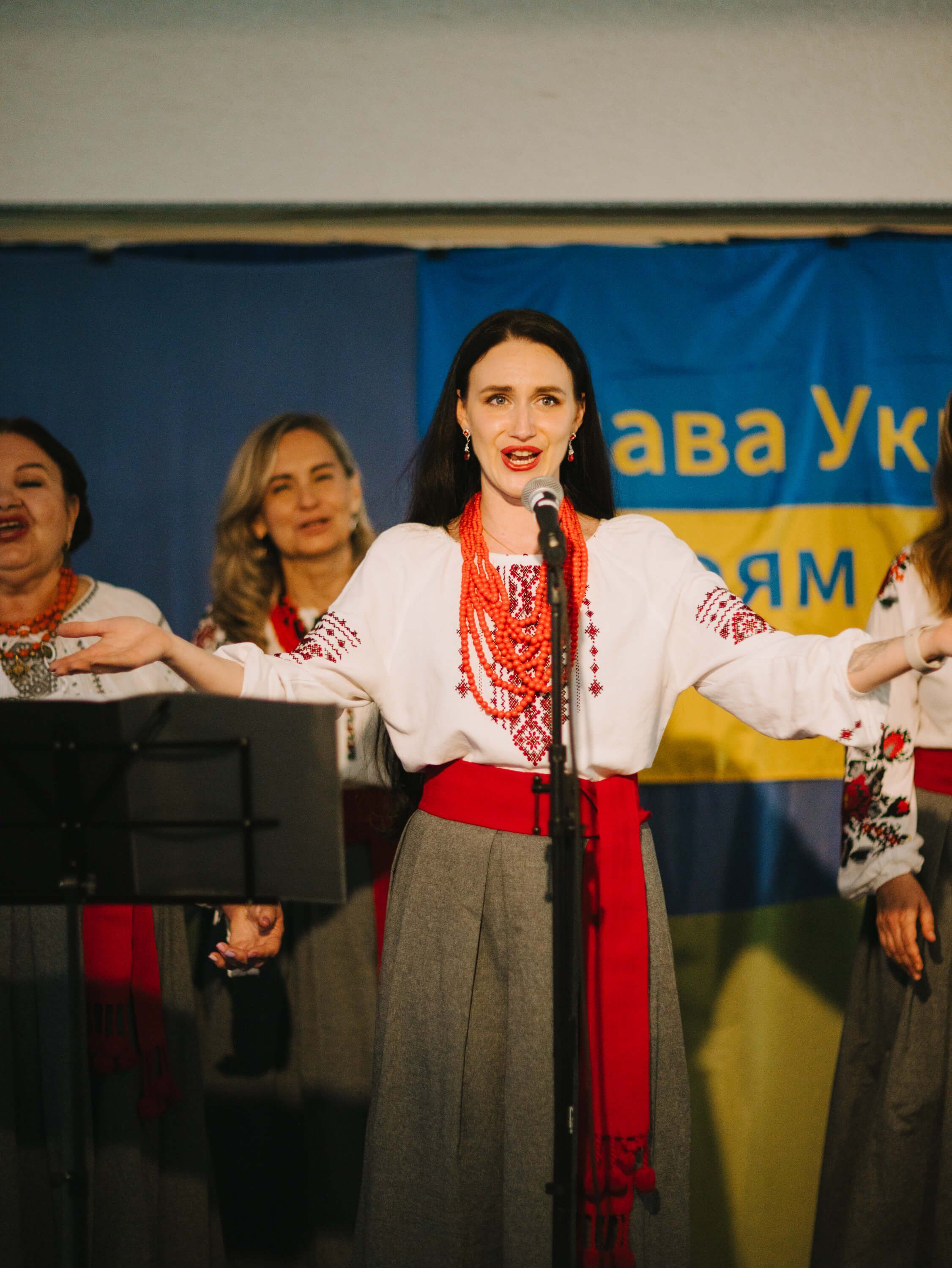 Woman in traditional Ukrainian embroidered dress and red beaded necklace speaks at microphone during fundraiser dinner event