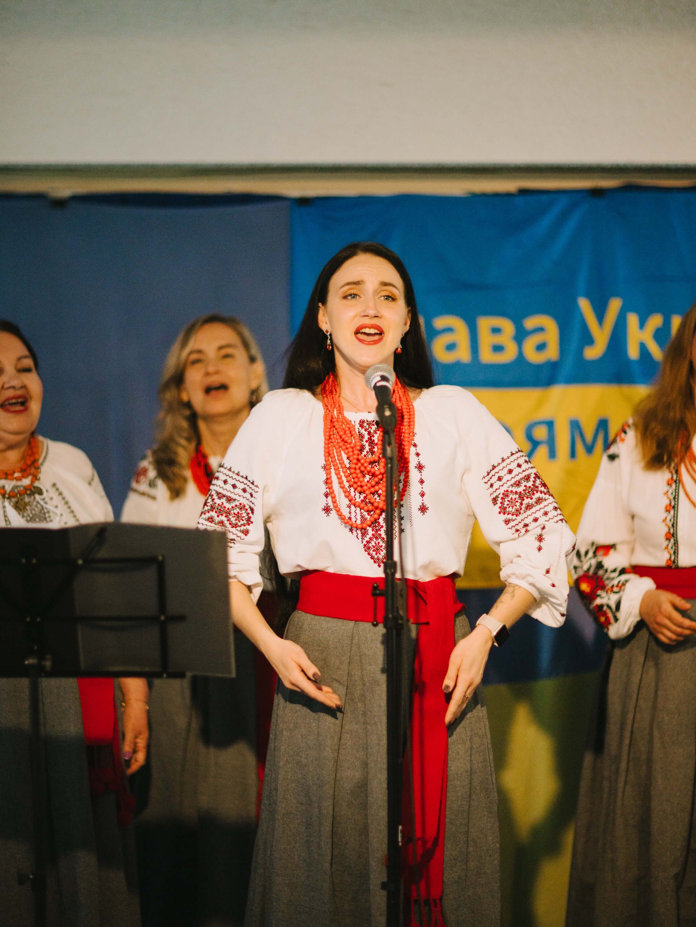 Woman in embroidered white shirt and red sash sings at microphone with three other performers in traditional Ukrainian dress behind her