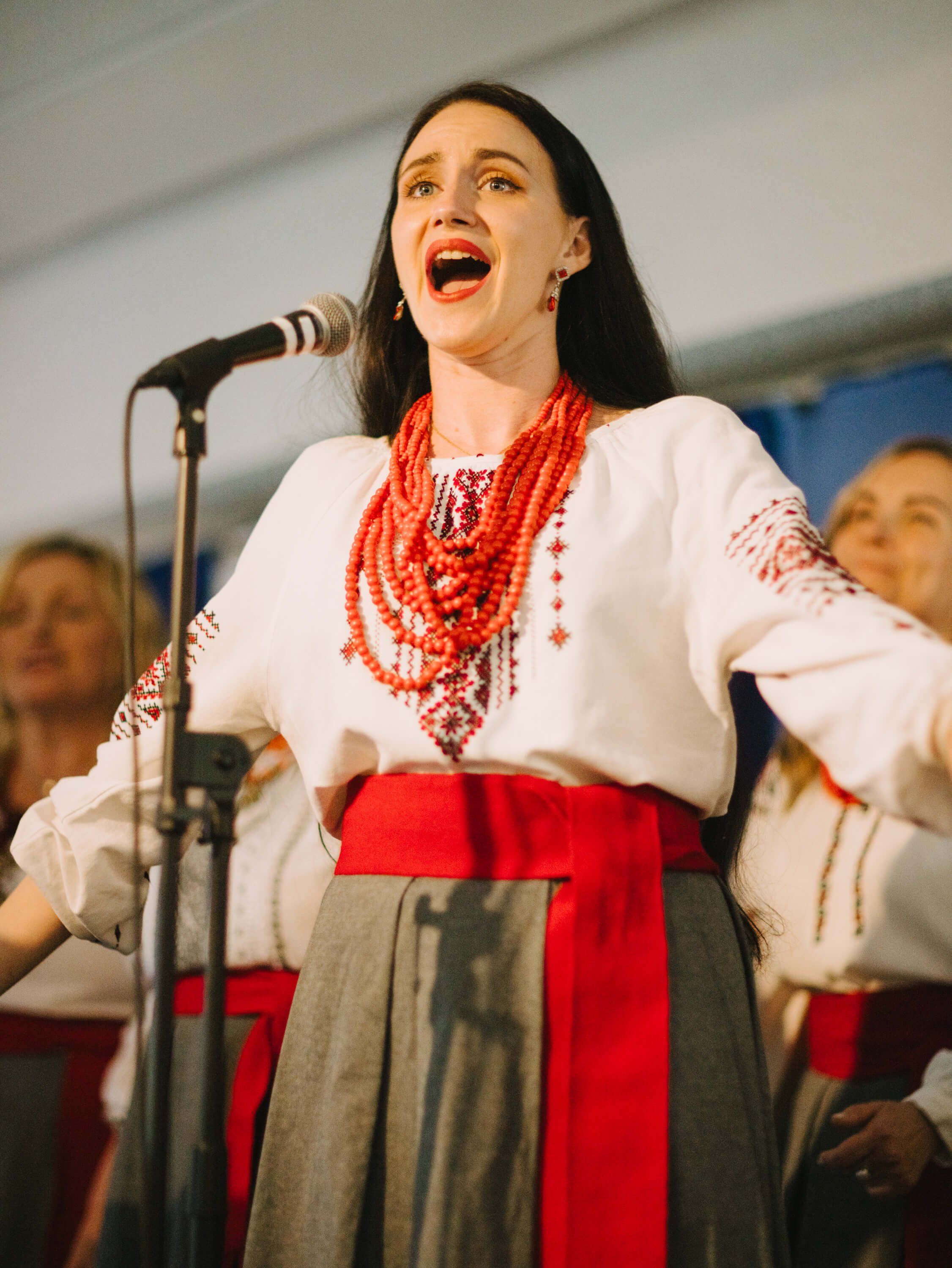 Woman in traditional Ukrainian embroidered white blouse and red skirt singing joyfully at microphone during fundraiser dinner
