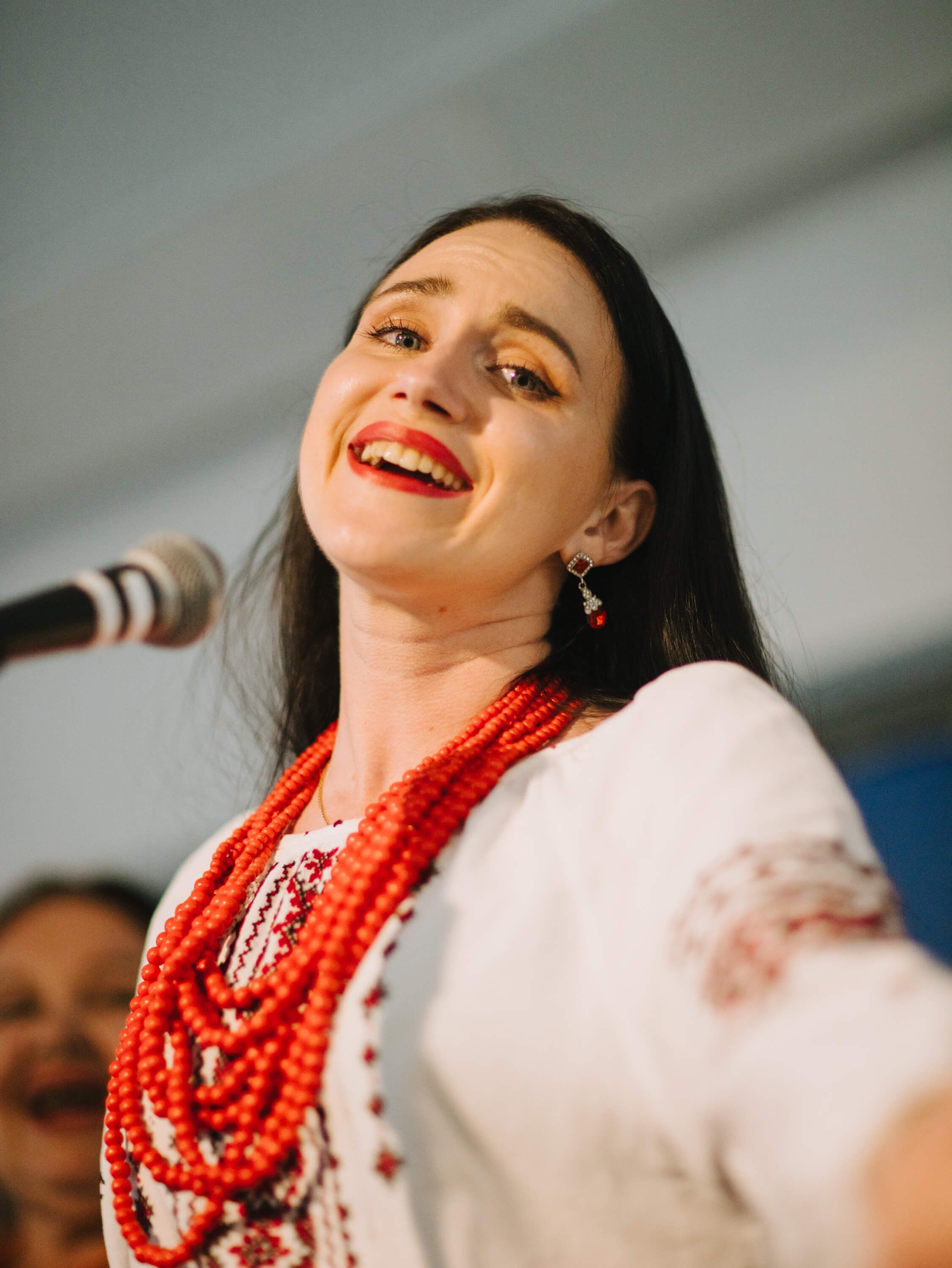 Woman in white embroidered shirt and red beaded necklace smiling at microphone during Ukrainian fundraiser dinner