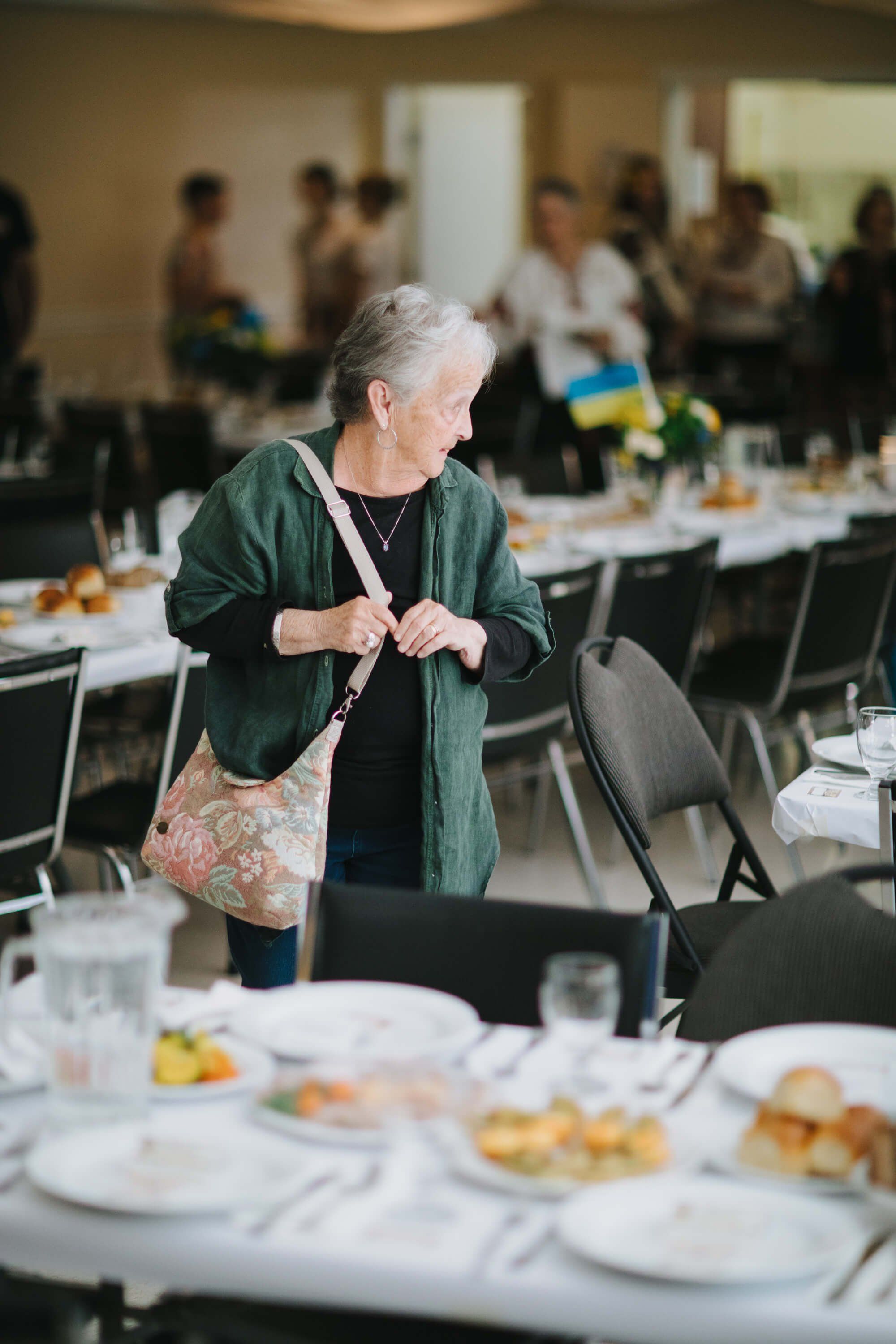 Woman in green cardigan stands at a dining table during the Ukrainian fundraiser dinner, with guests and place settings visible in the warm-lit hall behind her
