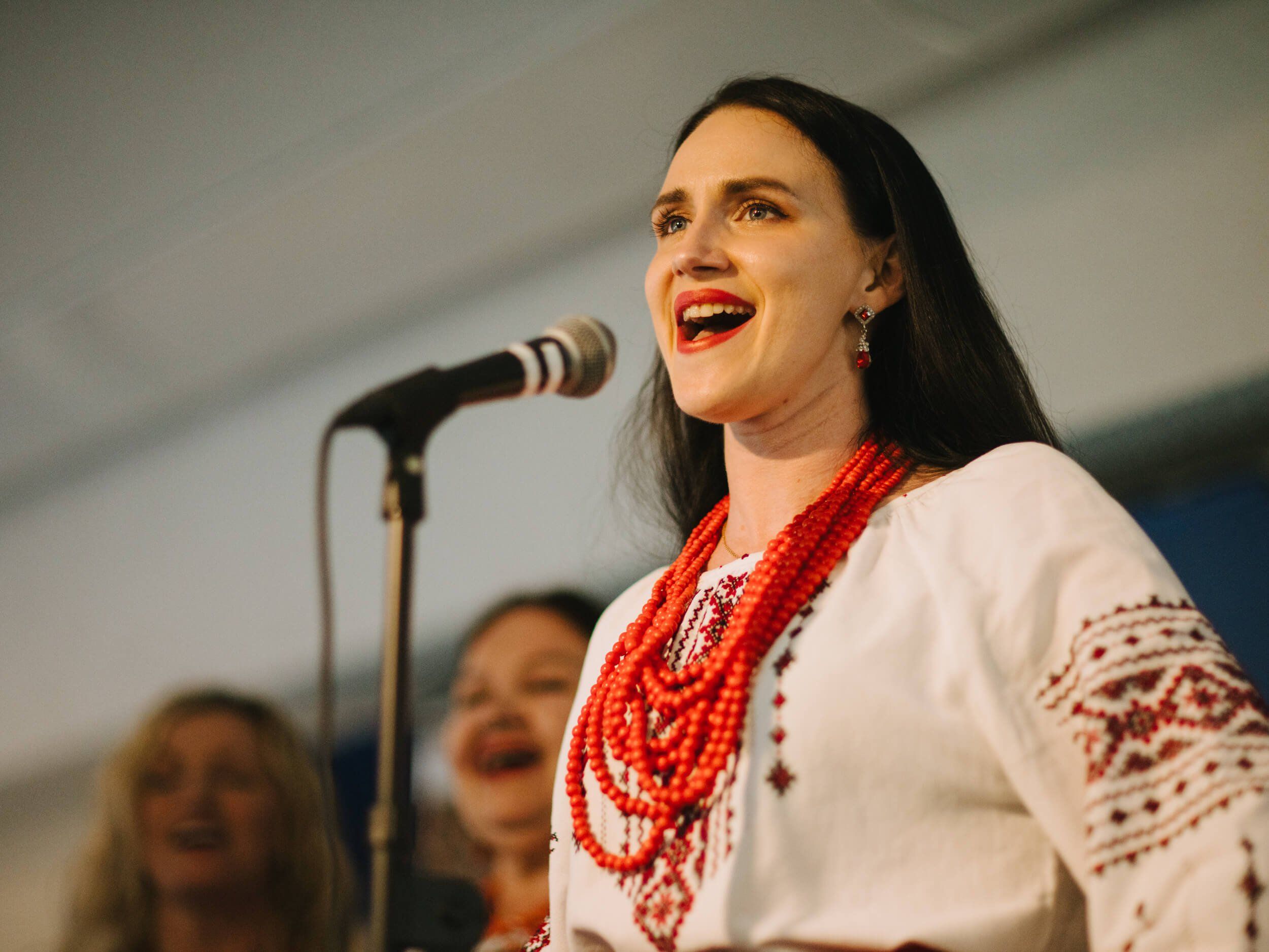Woman in embroidered white blouse and red beaded necklaces singing at microphone, smiling brightly during Ukrainian fundraiser dinner