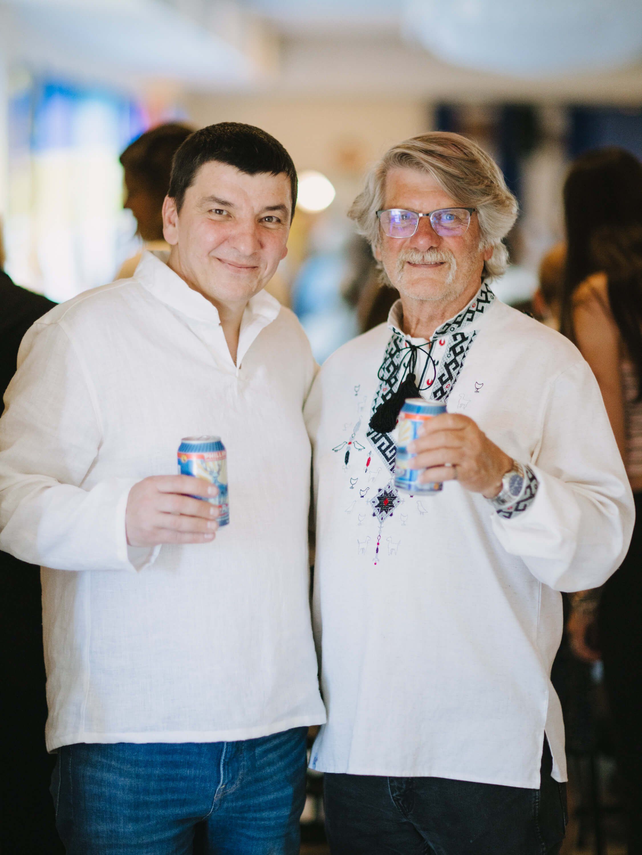 Two men in white shirts hold beverages and smile at a Ukrainian fundraiser dinner event indoors