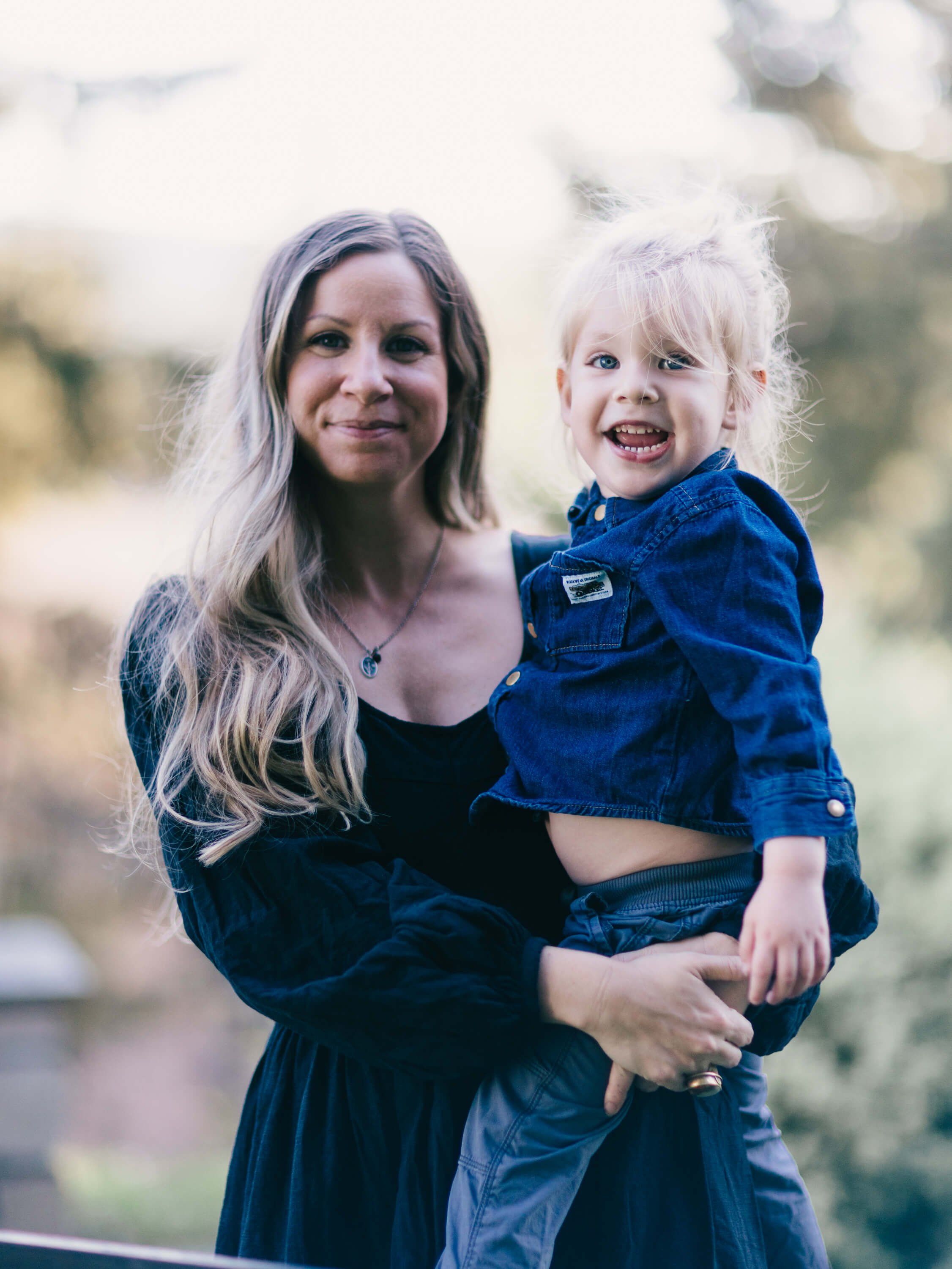 Woman in dark blue dress holding smiling blonde toddler in matching denim shirt, both gazing at camera with soft natural light filtering through blurred back…