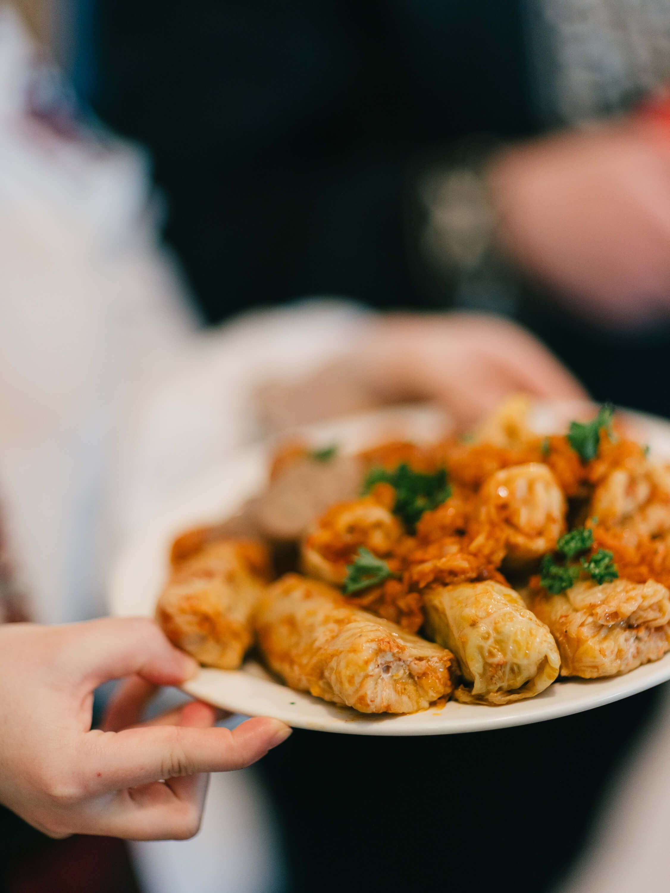 Hands holding a white plate of golden-brown Ukrainian pastries garnished with fresh herbs at an evening fundraiser dinner