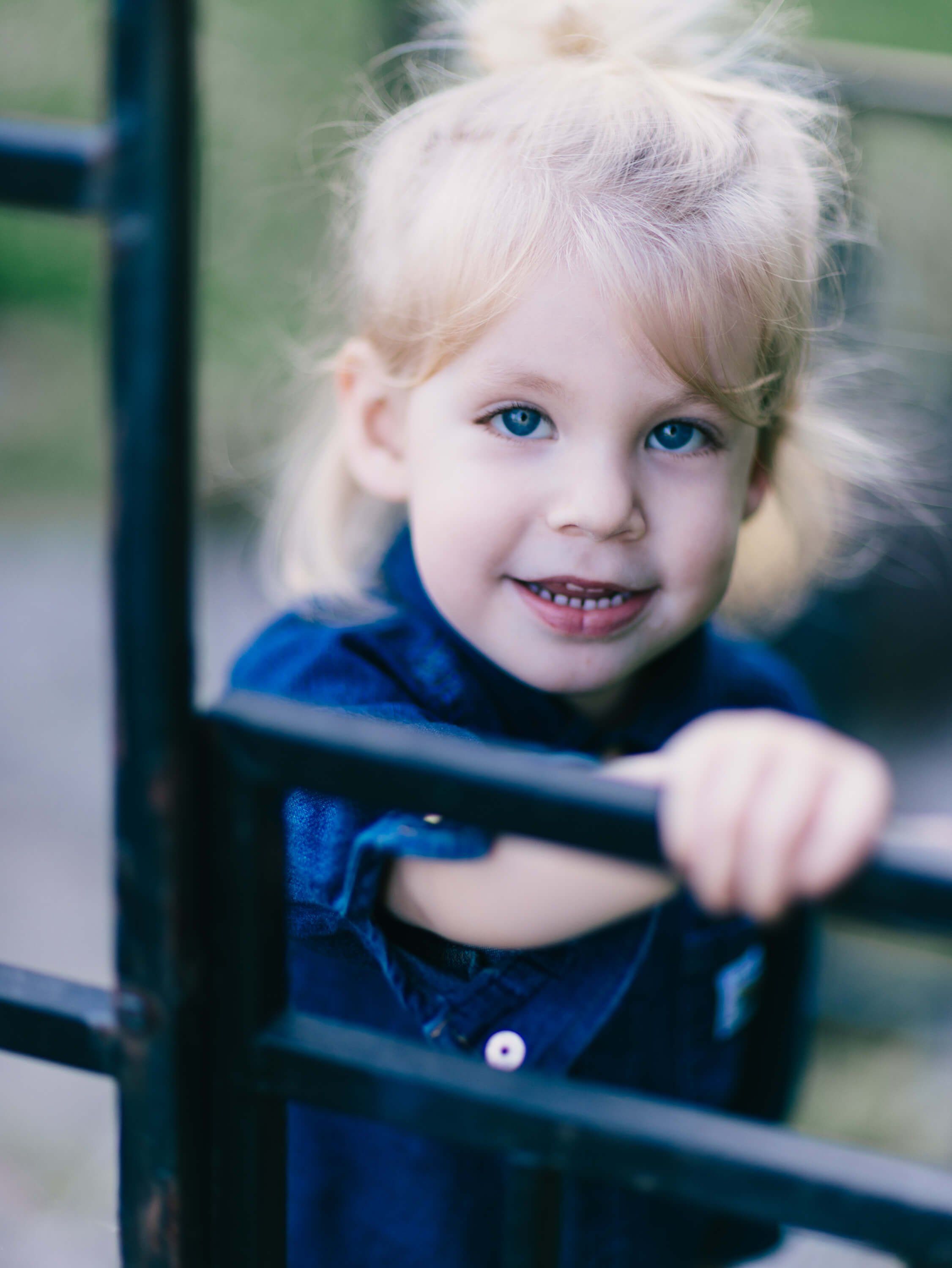 Young child with blonde hair smiling through a dark metal fence, wearing a blue jacket