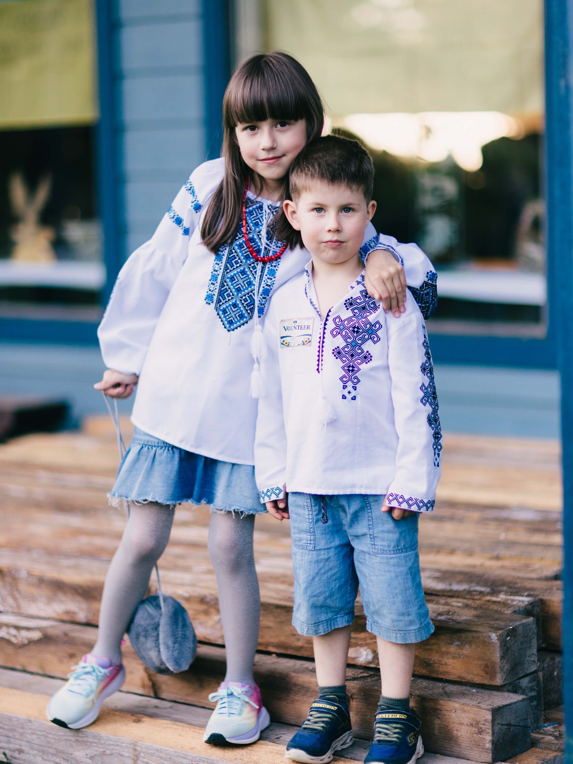 Two children in matching embroidered white shirts embrace outdoors on a wooden deck, blue buildings visible behind them