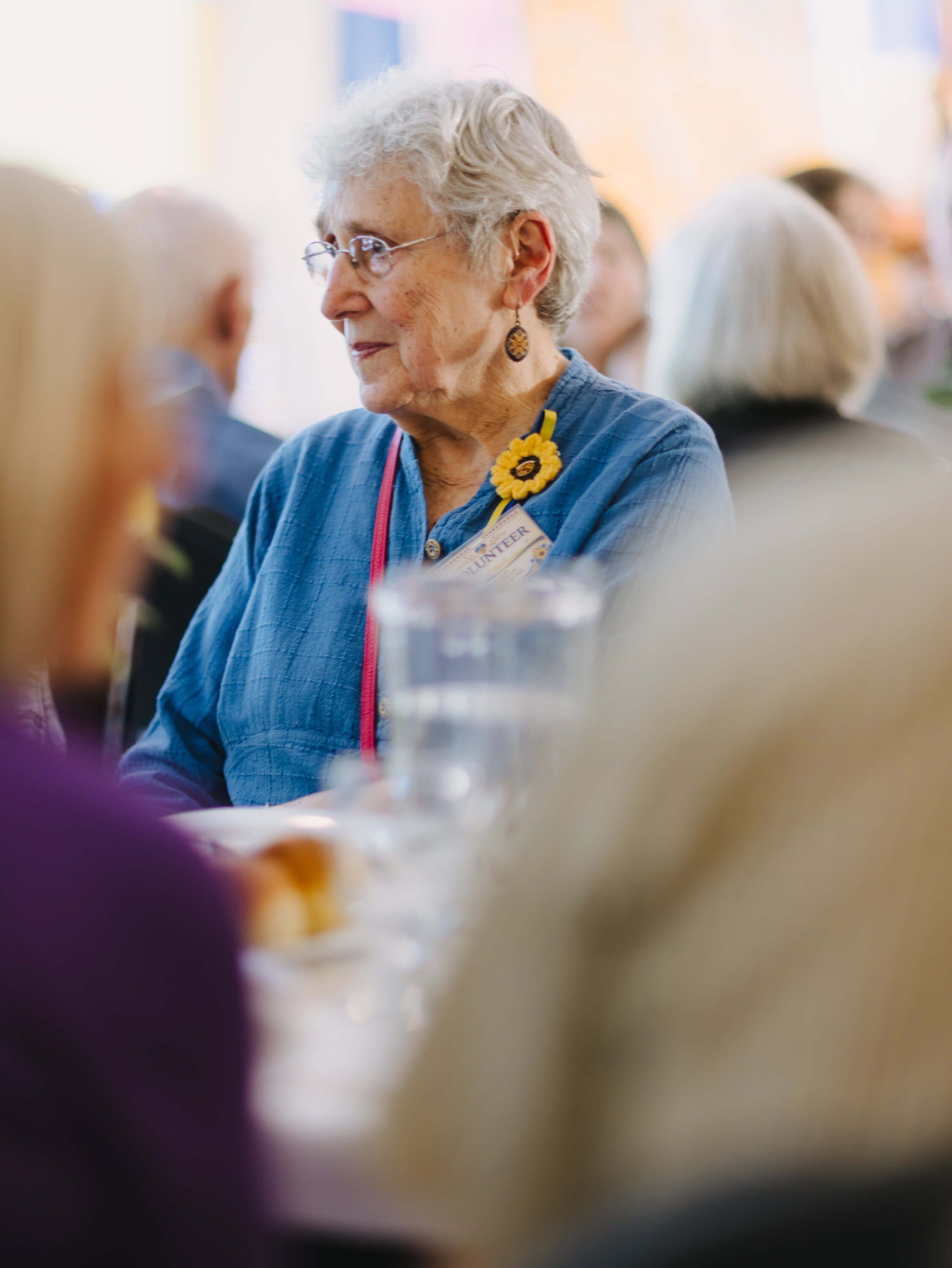 Senior woman in blue shirt and glasses smiles while conversing at a candlelit dinner table surrounded by guests