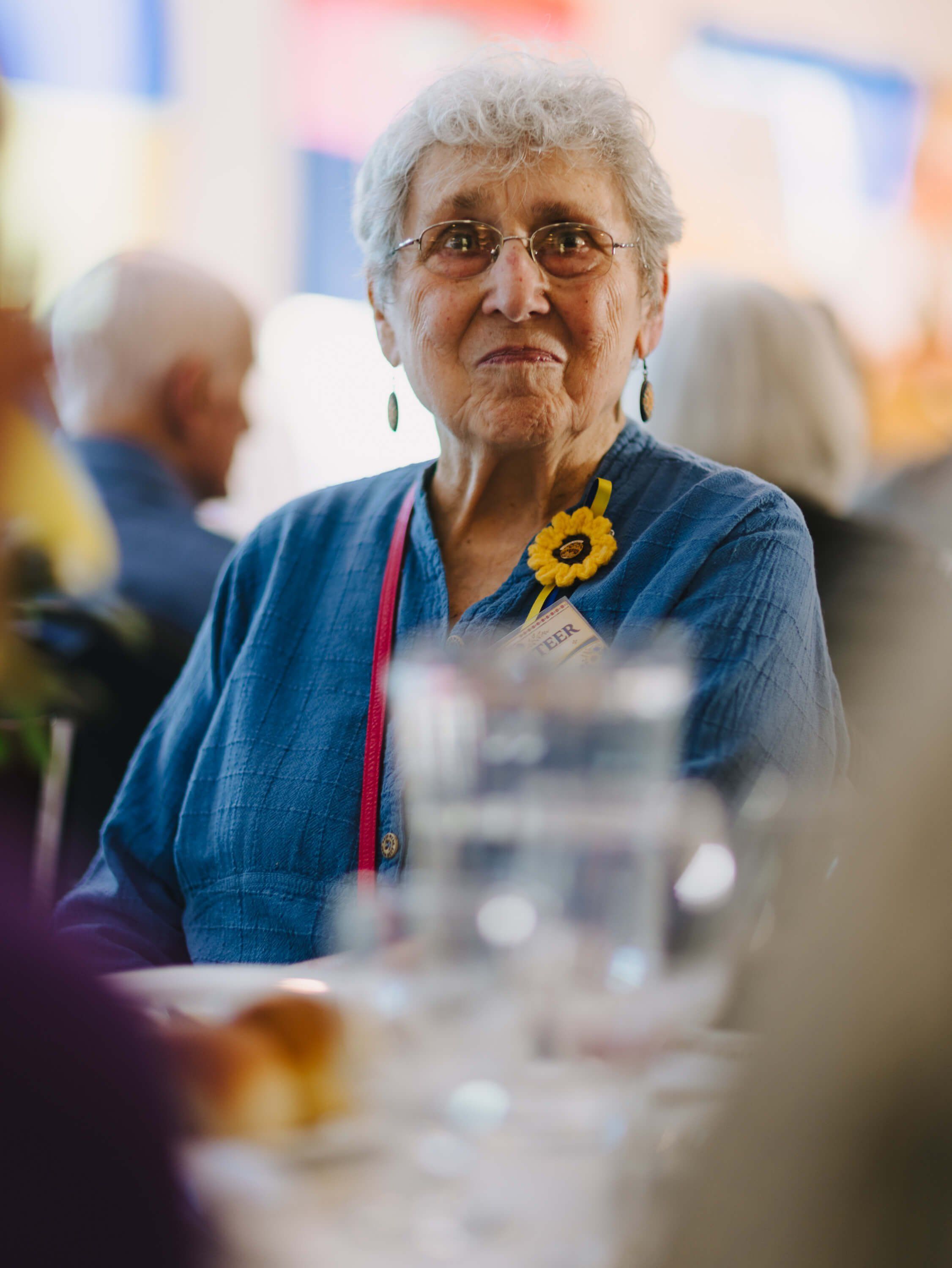 Elderly woman in blue shirt with sunflower pin at dinner table, warm indoor lighting and blurred background