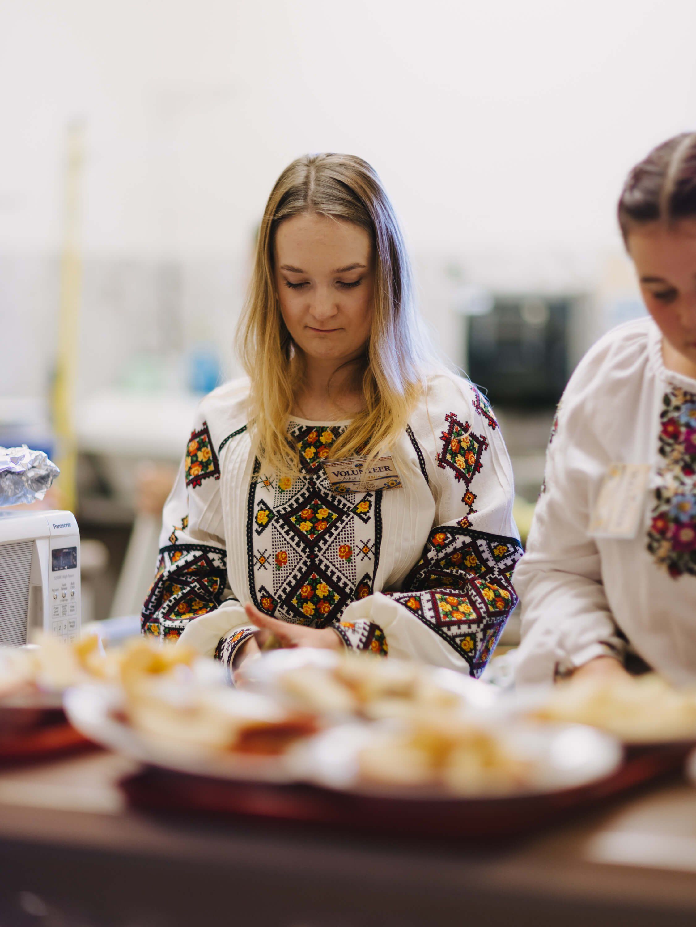 Woman in embroidered Ukrainian dress gazes down at plates of food during the Salt Spring Island fundraiser dinner