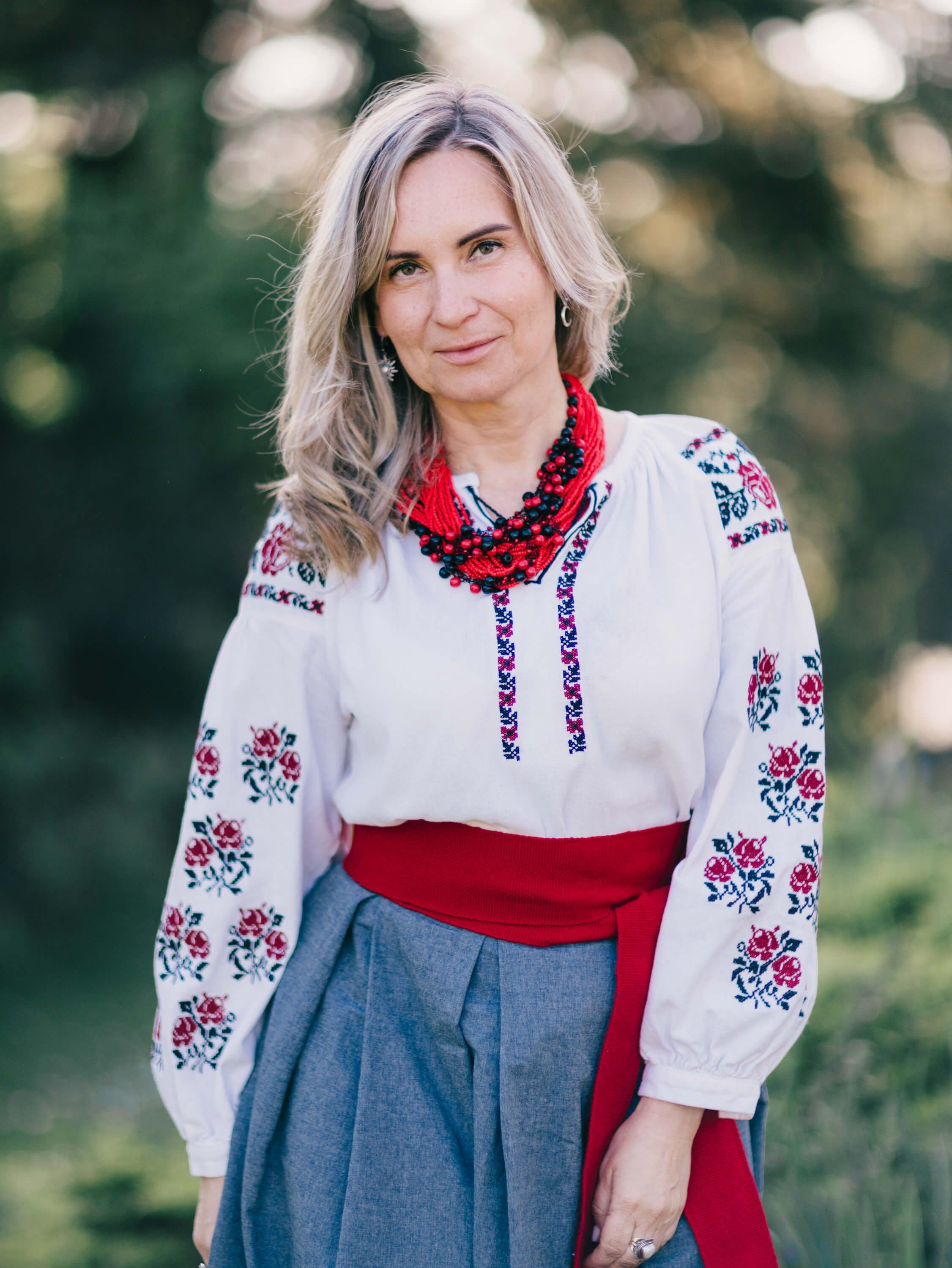 Woman in embroidered white linen blouse and red sash smiles at camera against soft-focus green backdrop