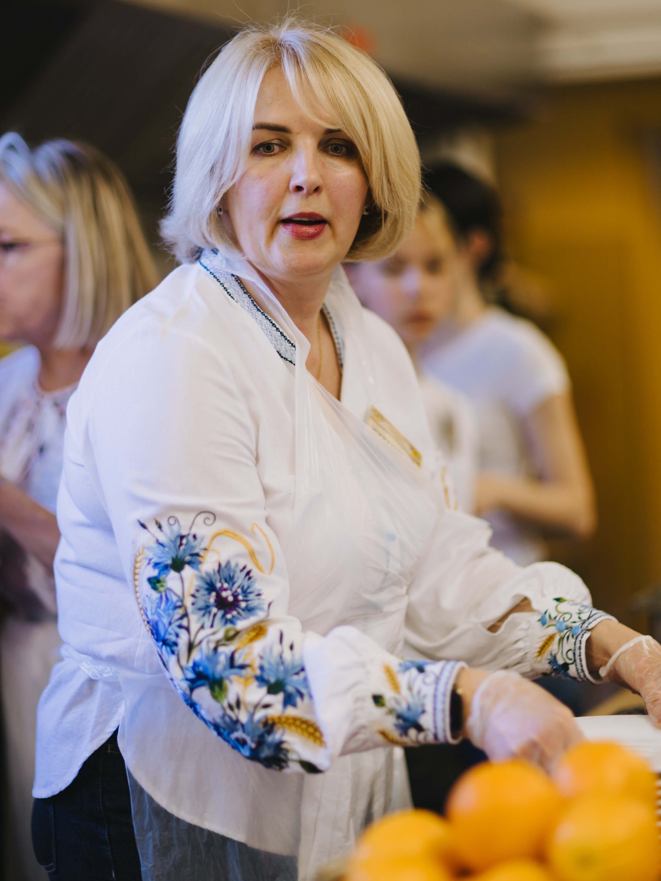 Woman in white embroidered blouse at Ukrainian fundraiser dinner, warm indoor lighting, fresh citrus visible in foreground