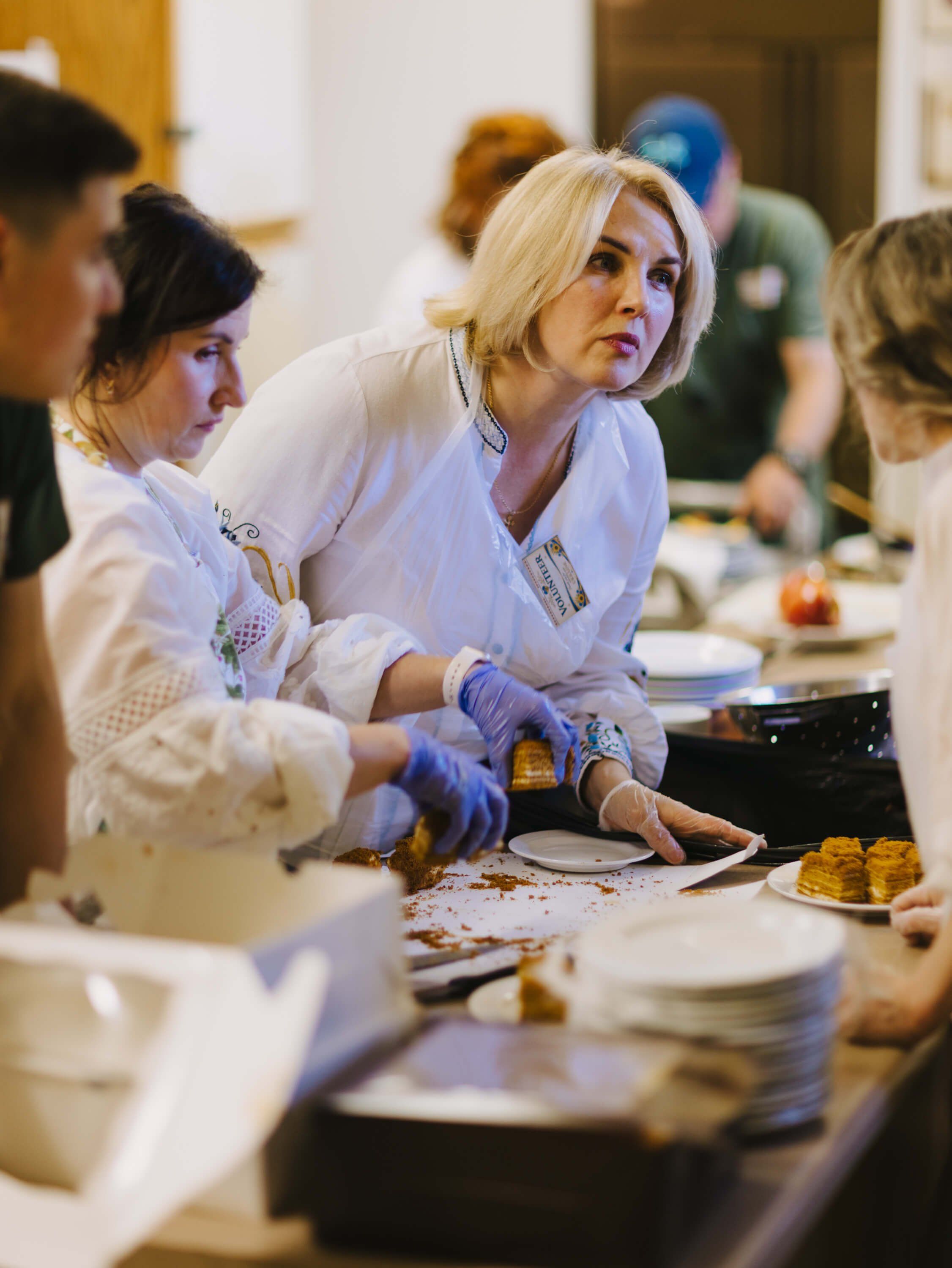 Chef in white apron and blue gloves plates food at a communal dinner table, focused on her work among gathered guests