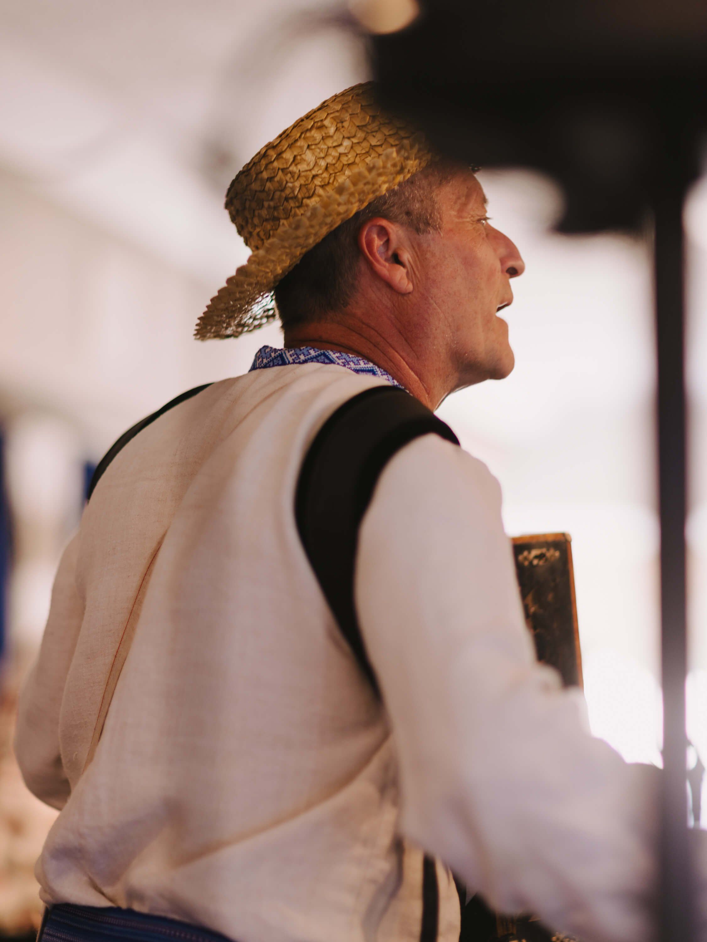 Man in cream vest and straw hat speaks animatedly at a candlelit evening event, warm ambient light highlighting the intimate atmosphere
