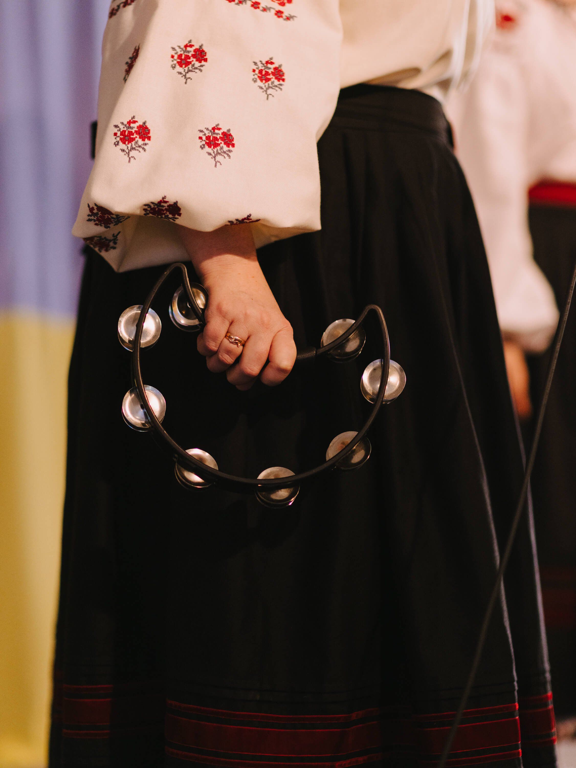 Person in embroidered white blouse and black skirt holds decorative metal rattle with bells at Ukrainian fundraiser dinner