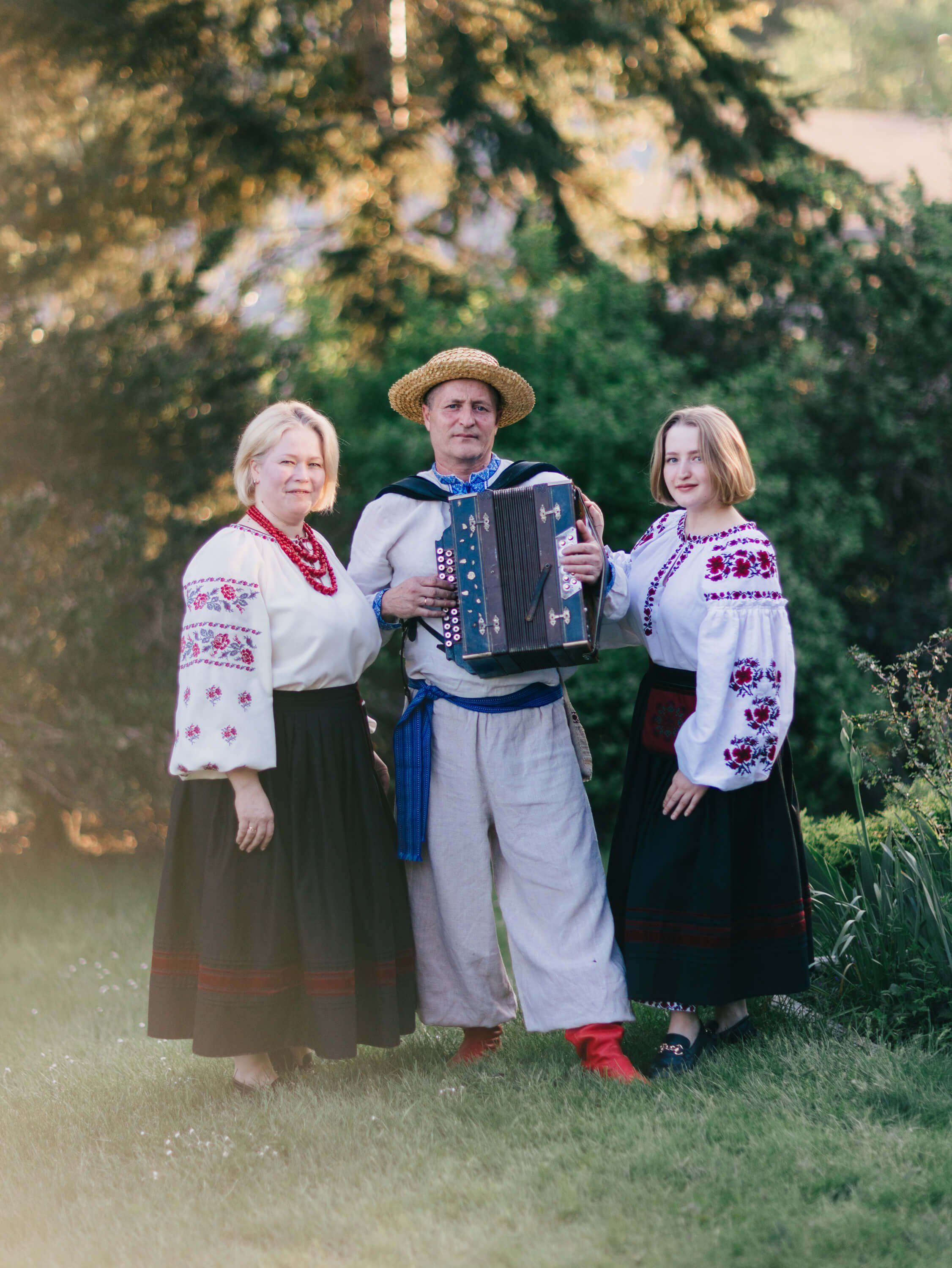 Three people in embroidered white and dark traditional Ukrainian clothing pose with an accordion beneath evergreen trees in golden evening light