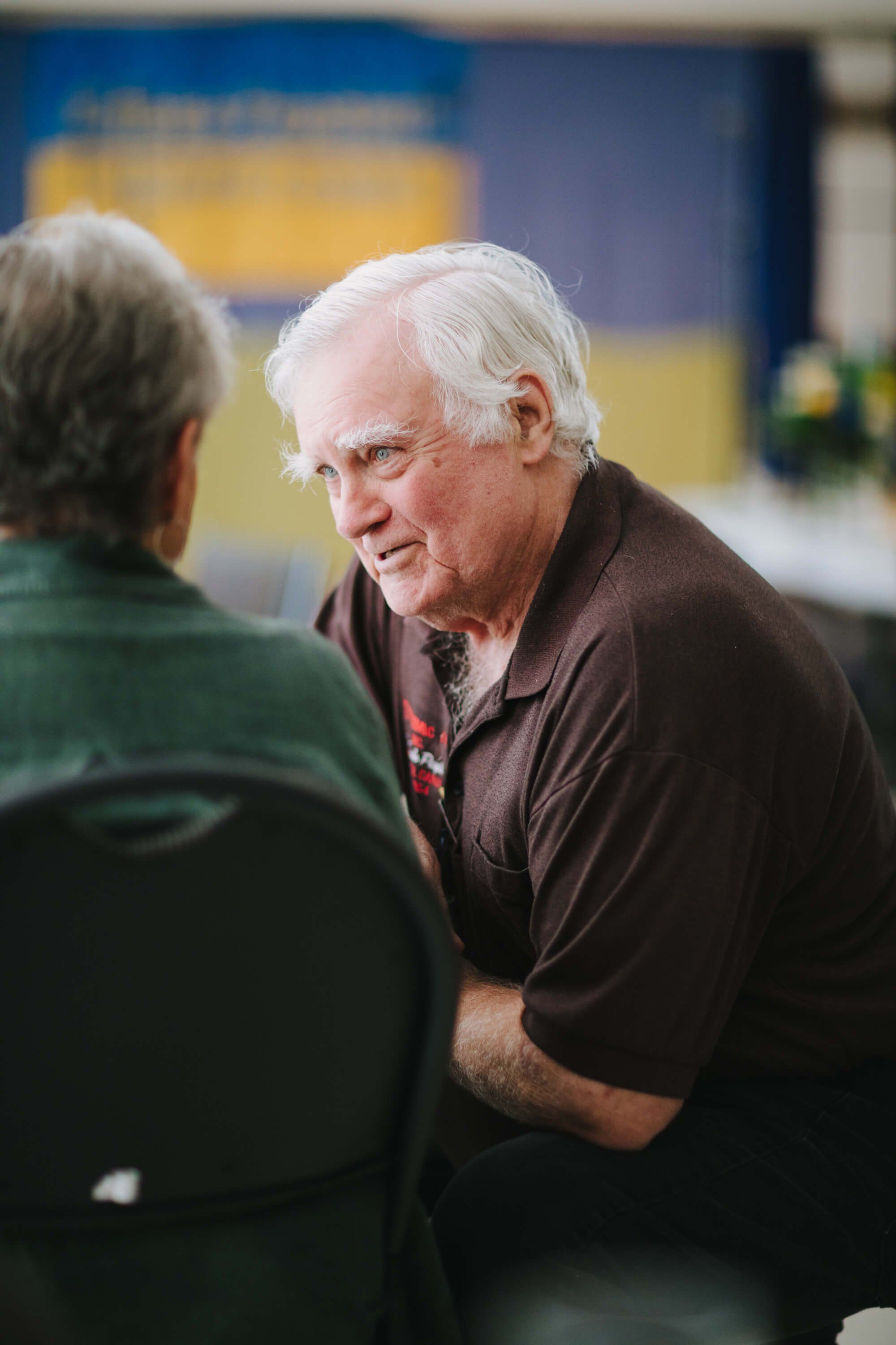 Two men converse at an indoor event, one wearing a dark polo shirt and glasses, smiling warmly during what appears to be a fundraiser dinner