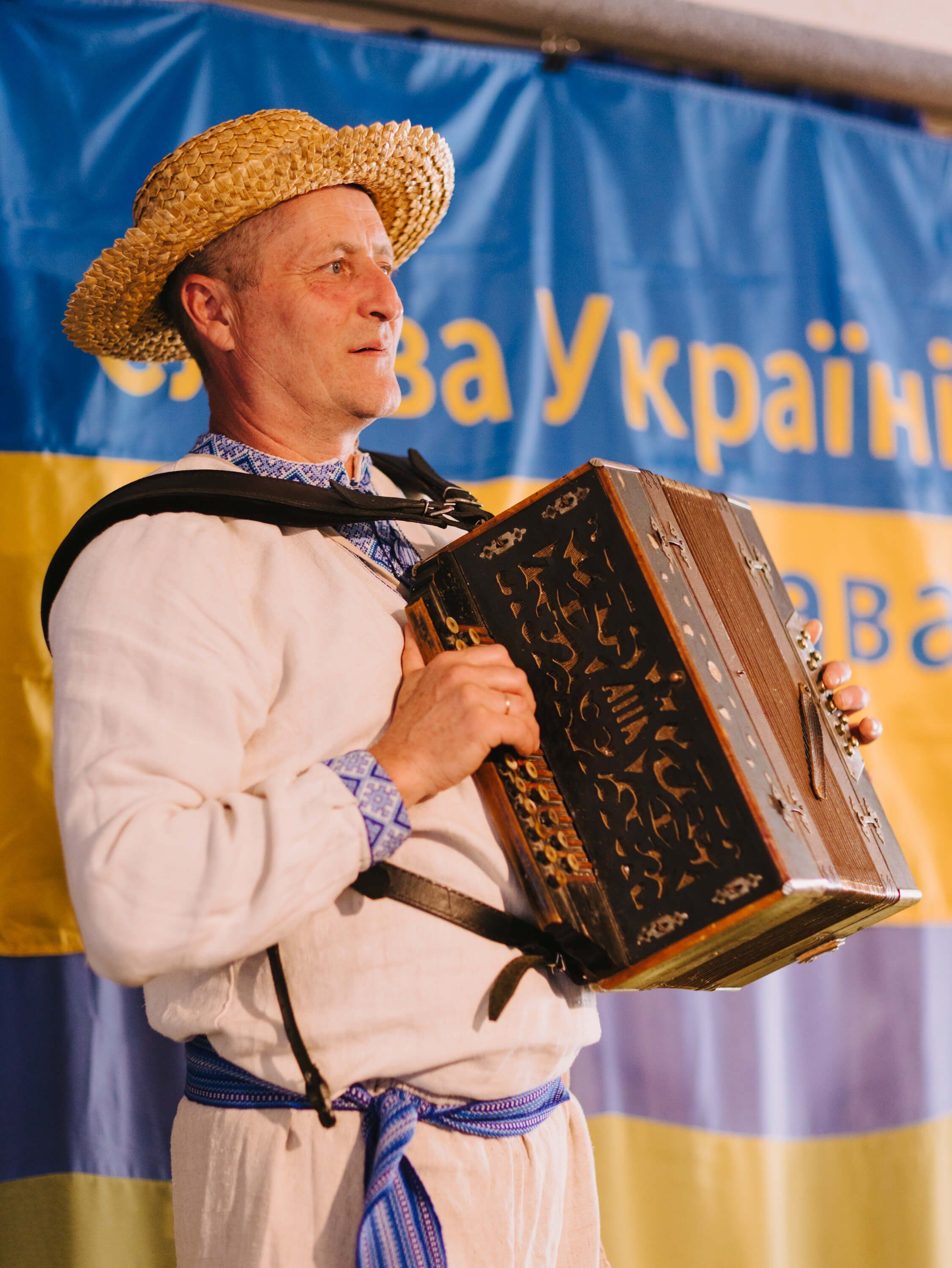 Man in traditional Ukrainian embroidered shirt and straw hat plays accordion on stage before blue and yellow backdrop