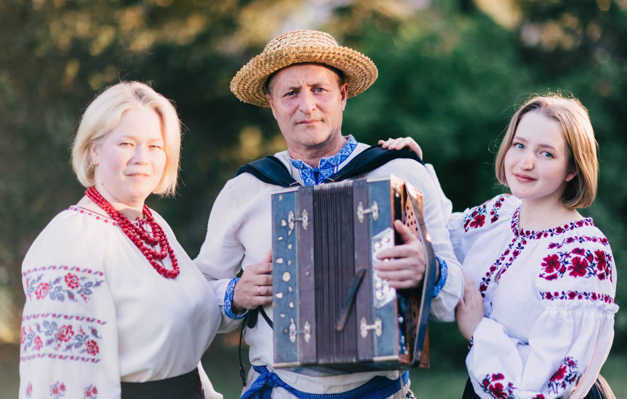 Three people in traditional Ukrainian embroidered shirts pose outdoors with an accordion, golden hour light filtering through trees behind them