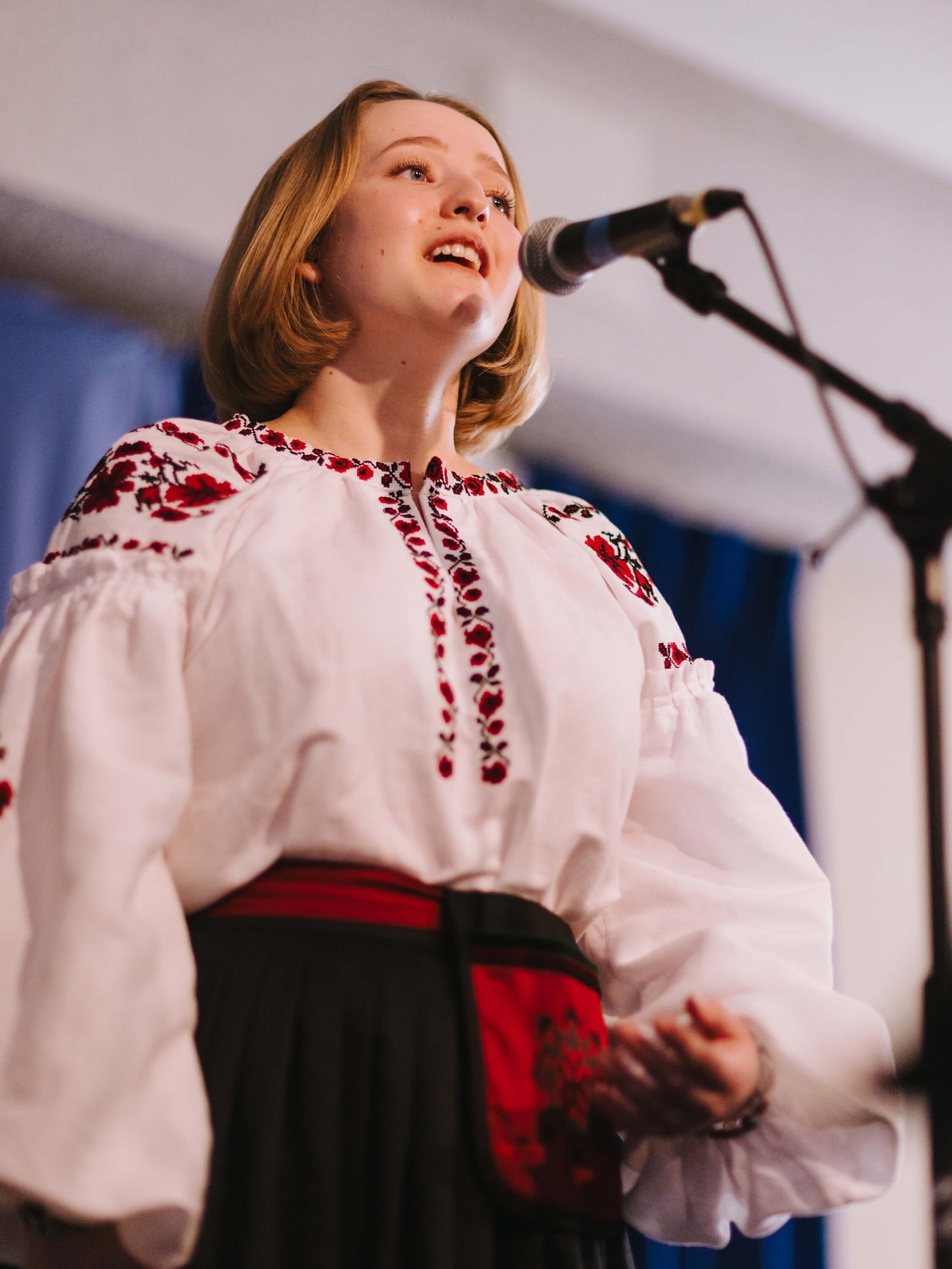 Woman in embroidered white linen shirt and red skirt singing at microphone, looking upward with warm expression