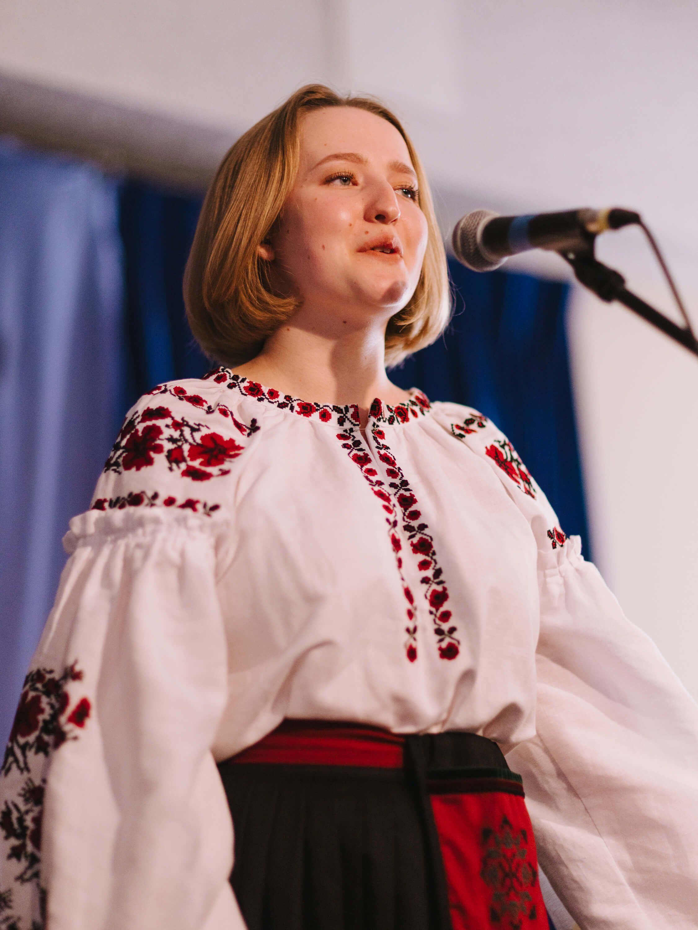 Woman in embroidered white and red Ukrainian dress singing at microphone, warm stage lighting casting soft shadows