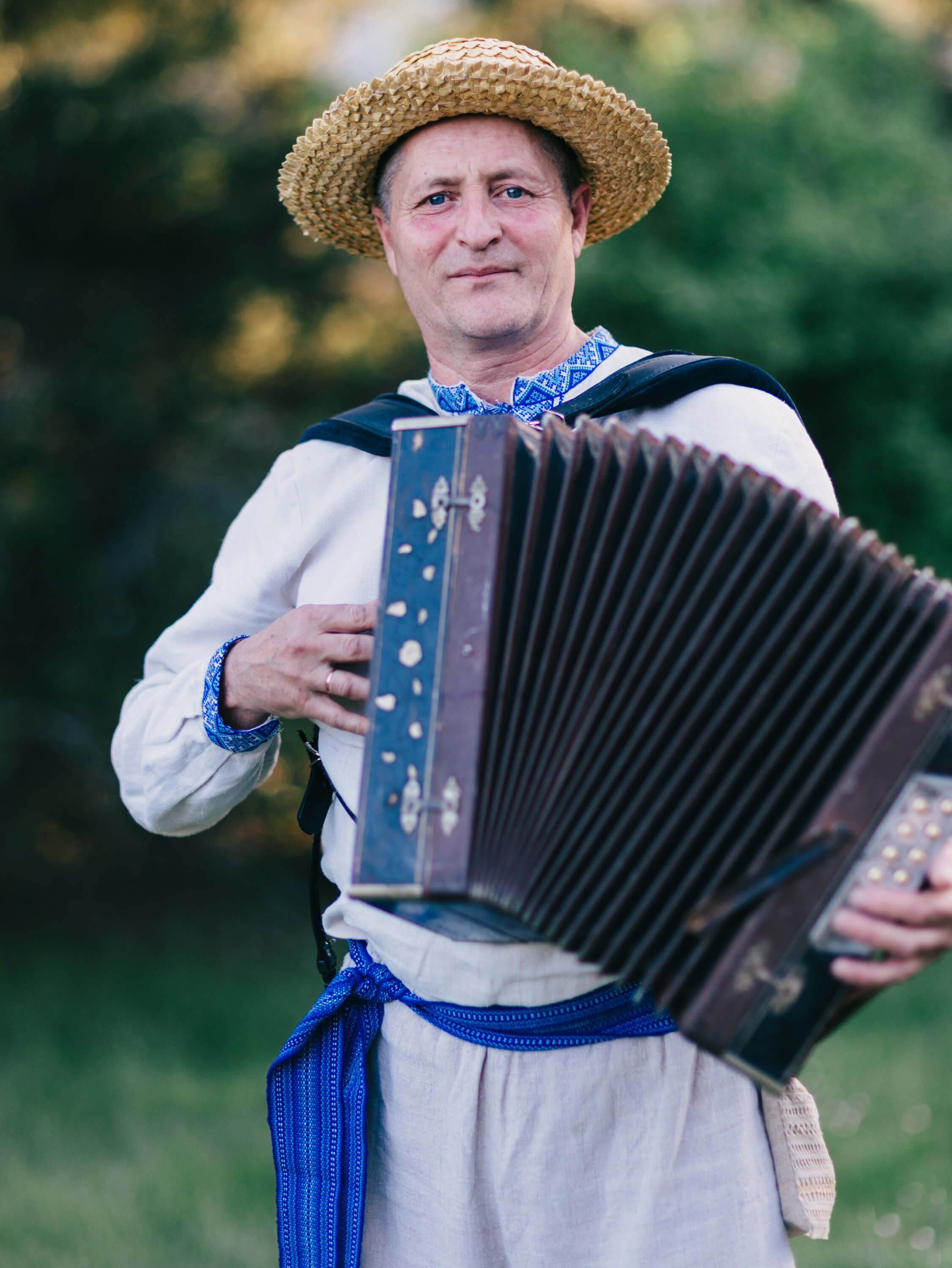 Man in embroidered linen shirt and straw hat plays accordion outdoors, wearing traditional blue sash and neckerchief