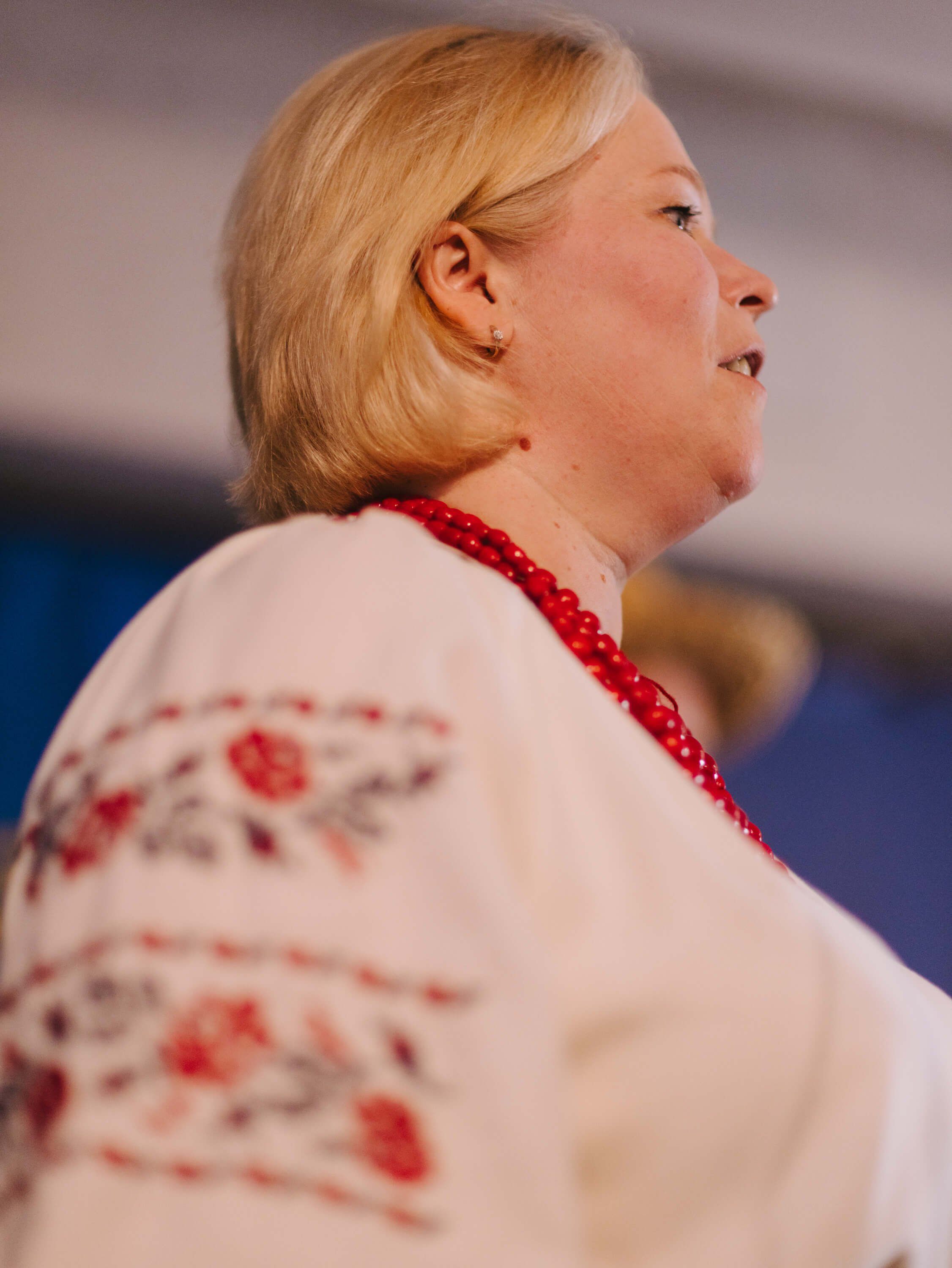 Woman in profile wearing embroidered white linen with red floral details, soft blonde hair, warm indoor lighting