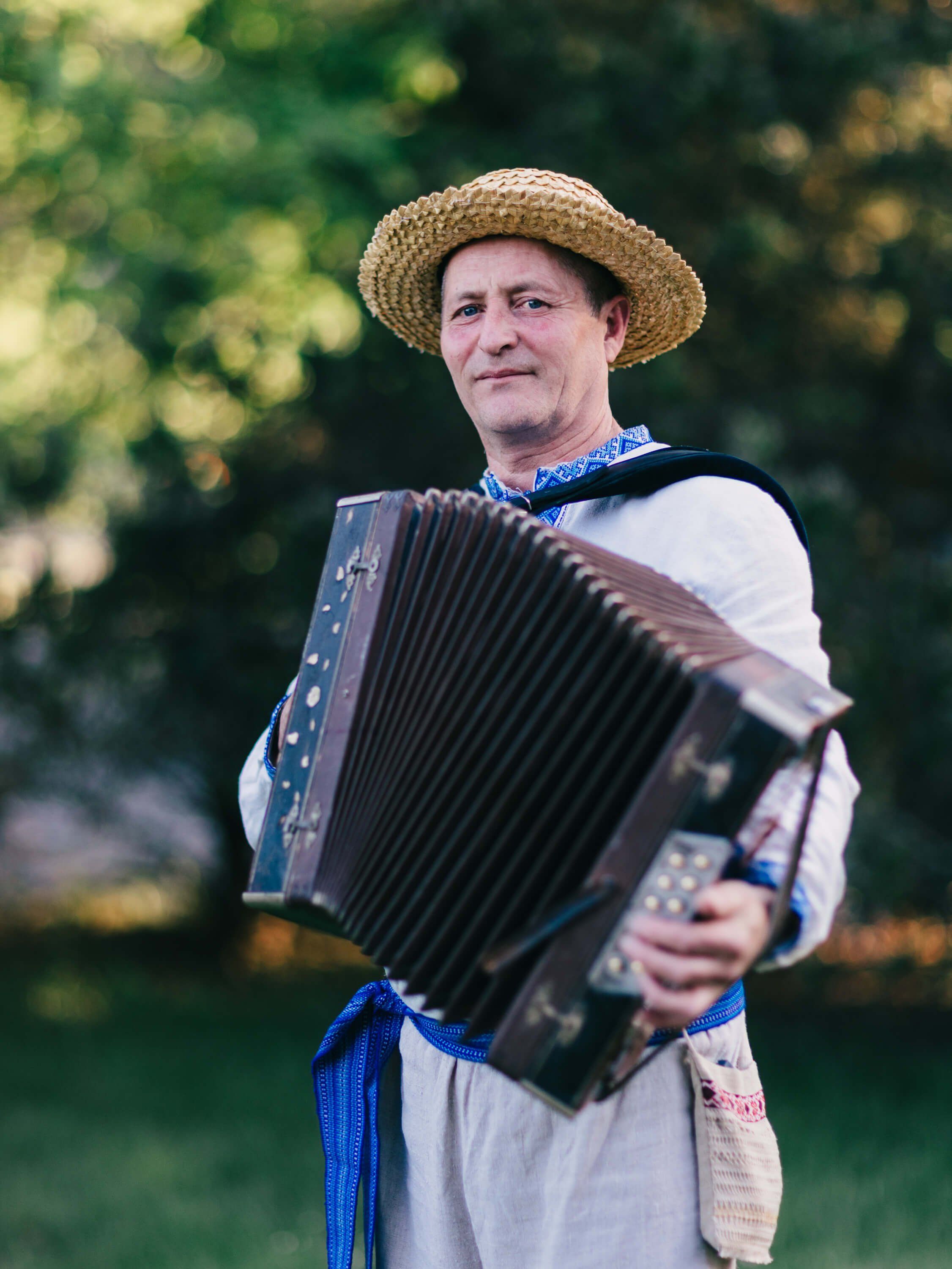 Man in straw hat and embroidered linen shirt plays accordion outdoors at sunset with green foliage behind