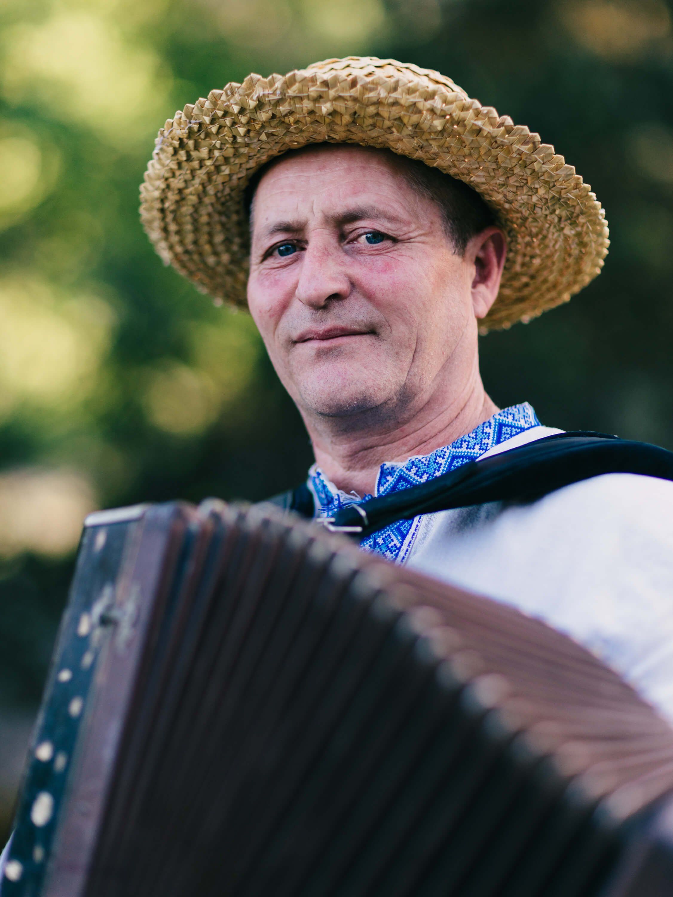 Musician in straw hat and blue bandana playing accordion outdoors, warm golden light filtering through green foliage behind