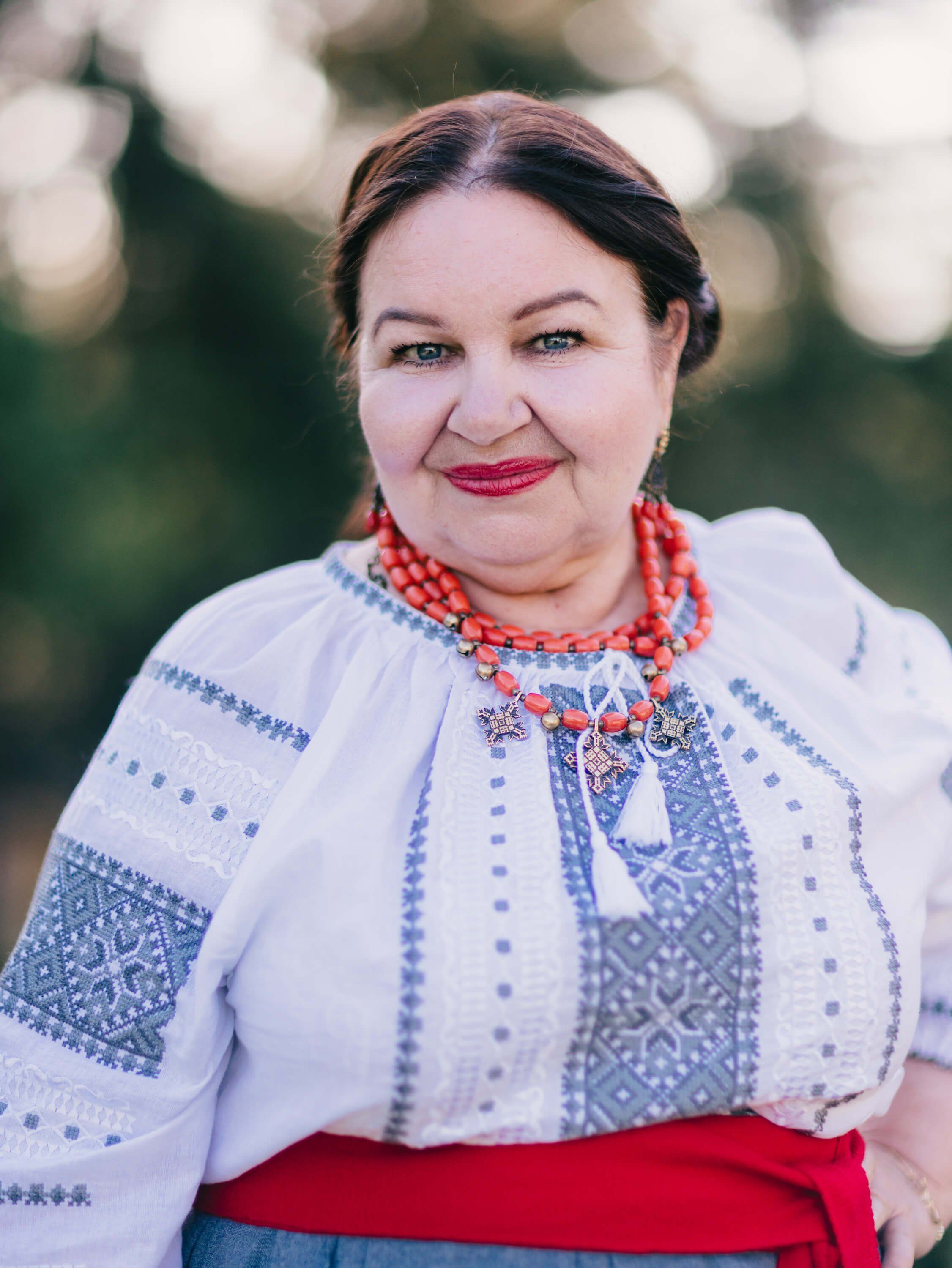 Woman in traditional Ukrainian embroidered white linen shirt and red skirt smiles at camera with blurred green foliage behind
