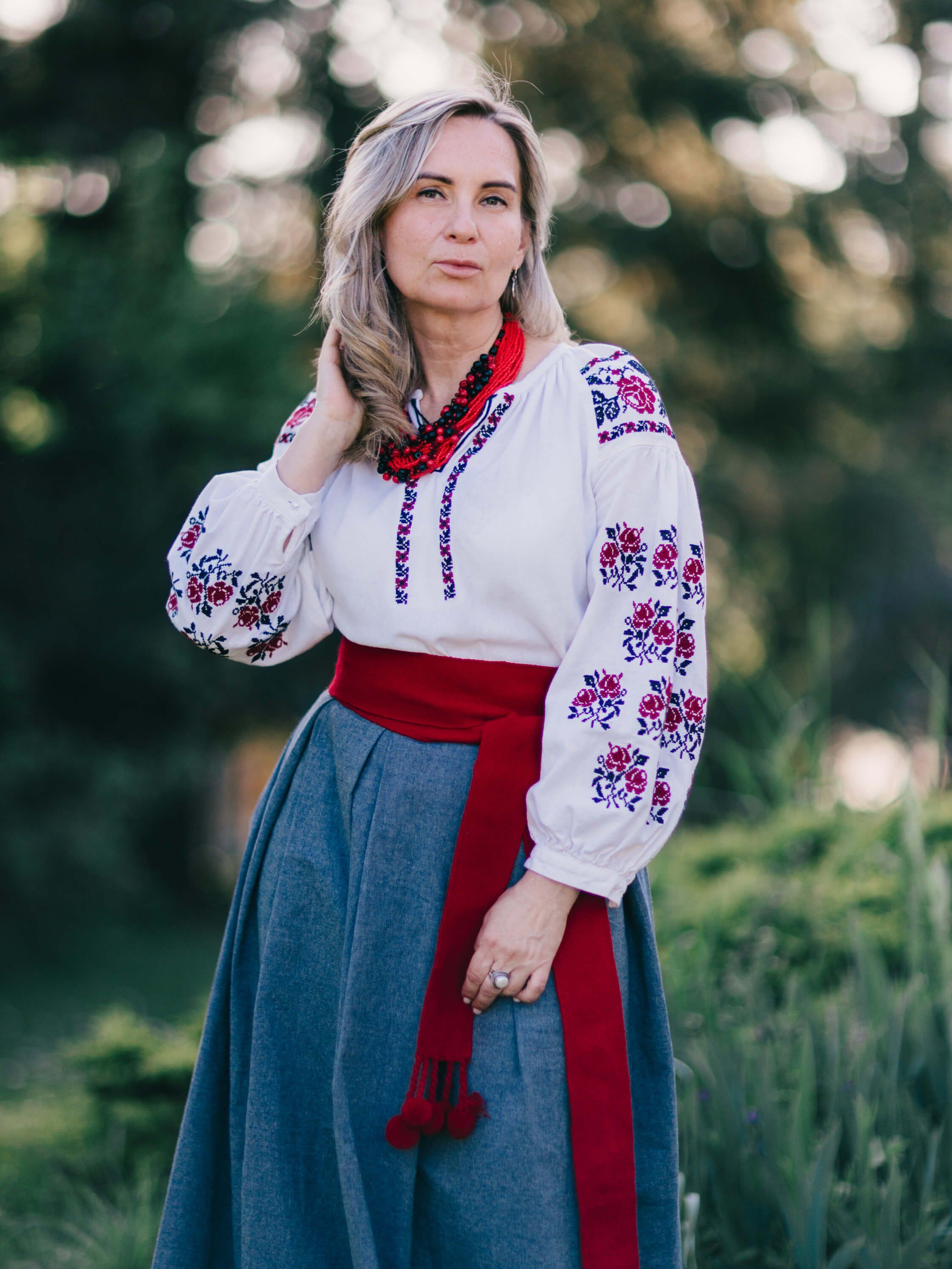 Woman in traditional Ukrainian embroidered blouse and red skirt poses outdoors at golden hour with soft forest backdrop