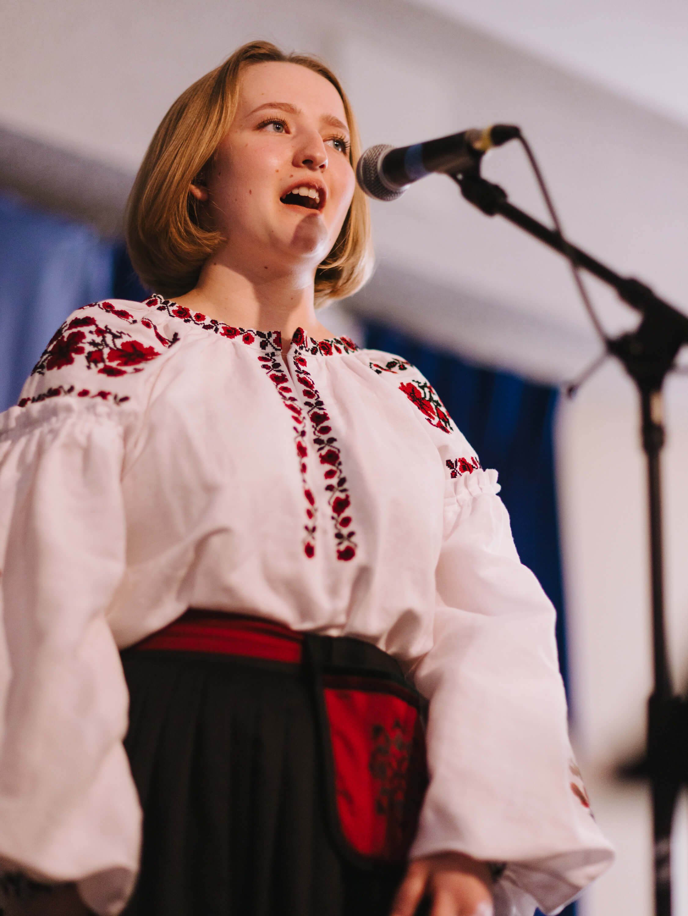 Woman in embroidered white linen shirt and red-black vest singing at microphone during Ukrainian fundraiser dinner