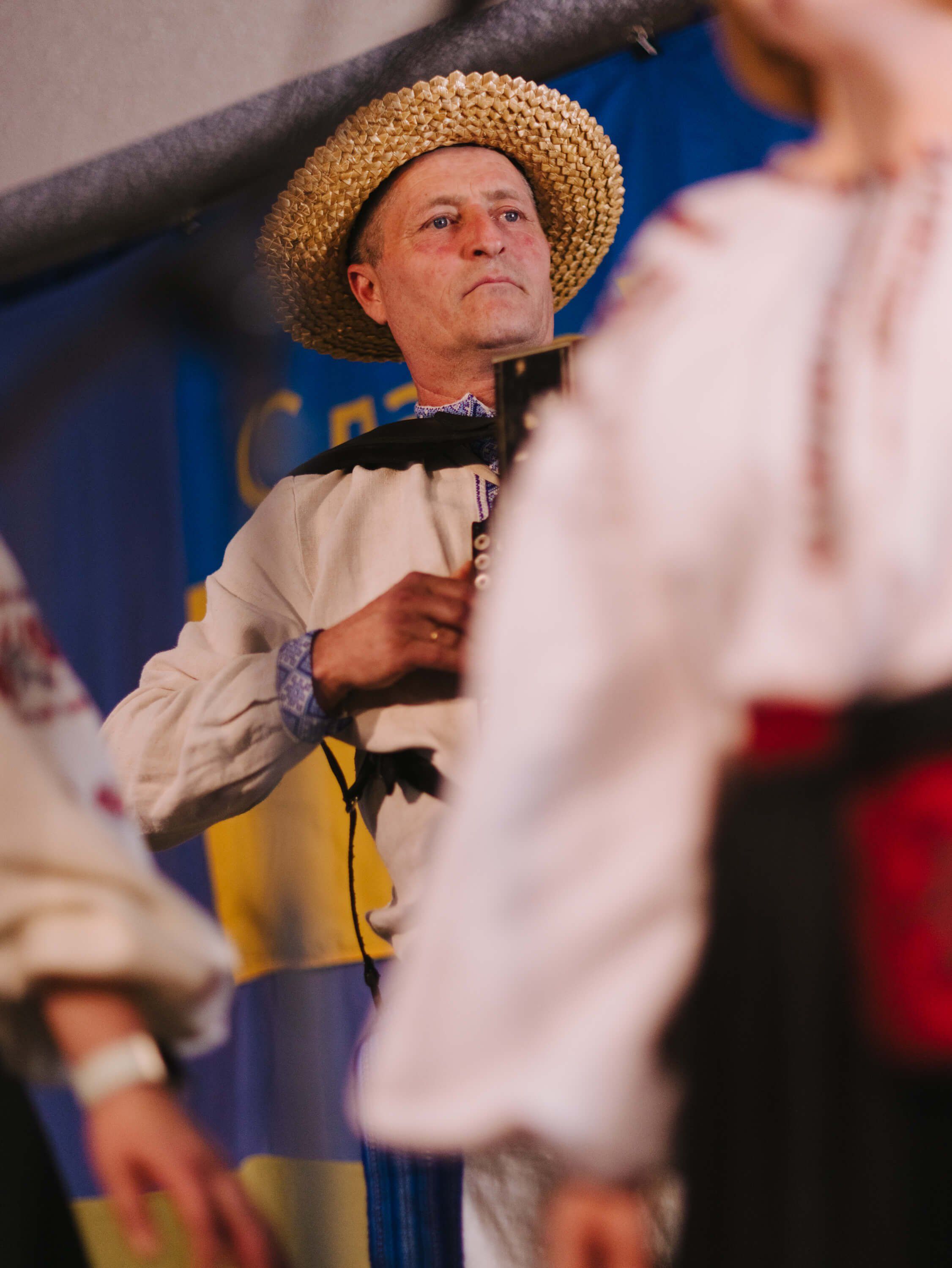 Man in straw hat and cream shirt holds microphone at indoor fundraiser dinner, warm lighting casting gentle shadows
