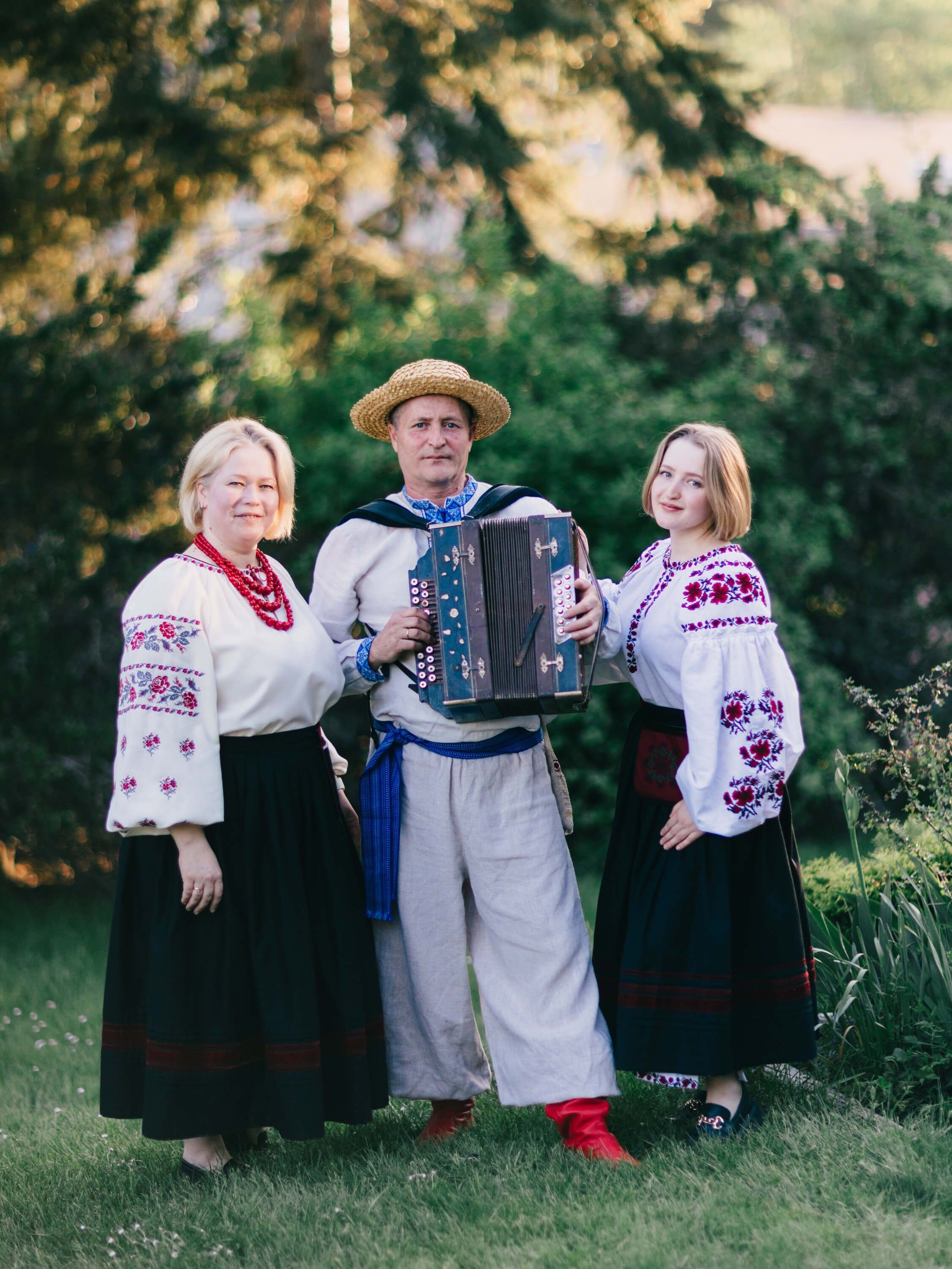 Three people in embroidered Ukrainian folk costumes stand together outdoors, the man in the center holding an accordion, golden sunlight filtering through tr…