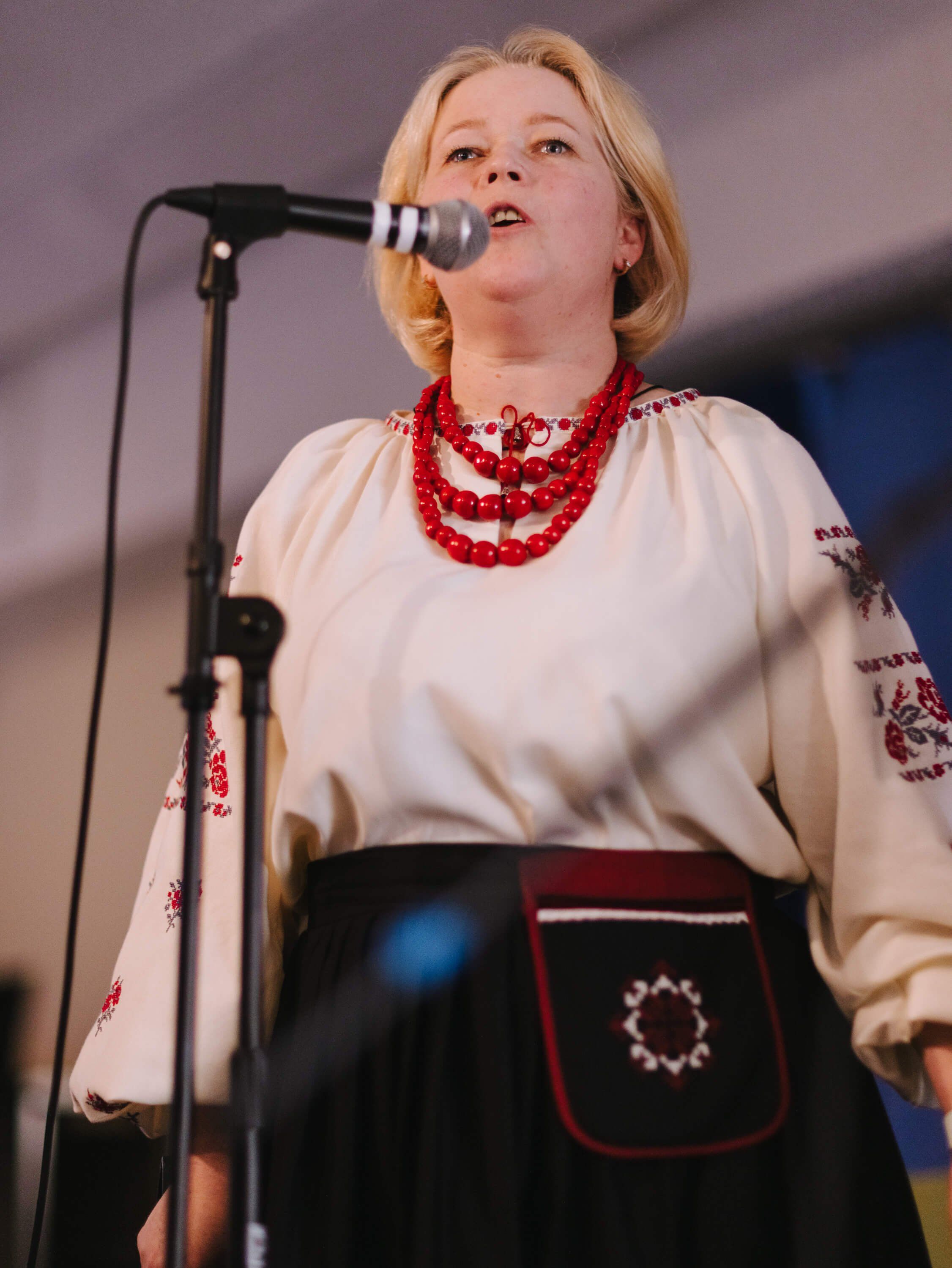 Blonde woman in embroidered Ukrainian blouse and red beaded necklace singing at microphone during fundraiser dinner