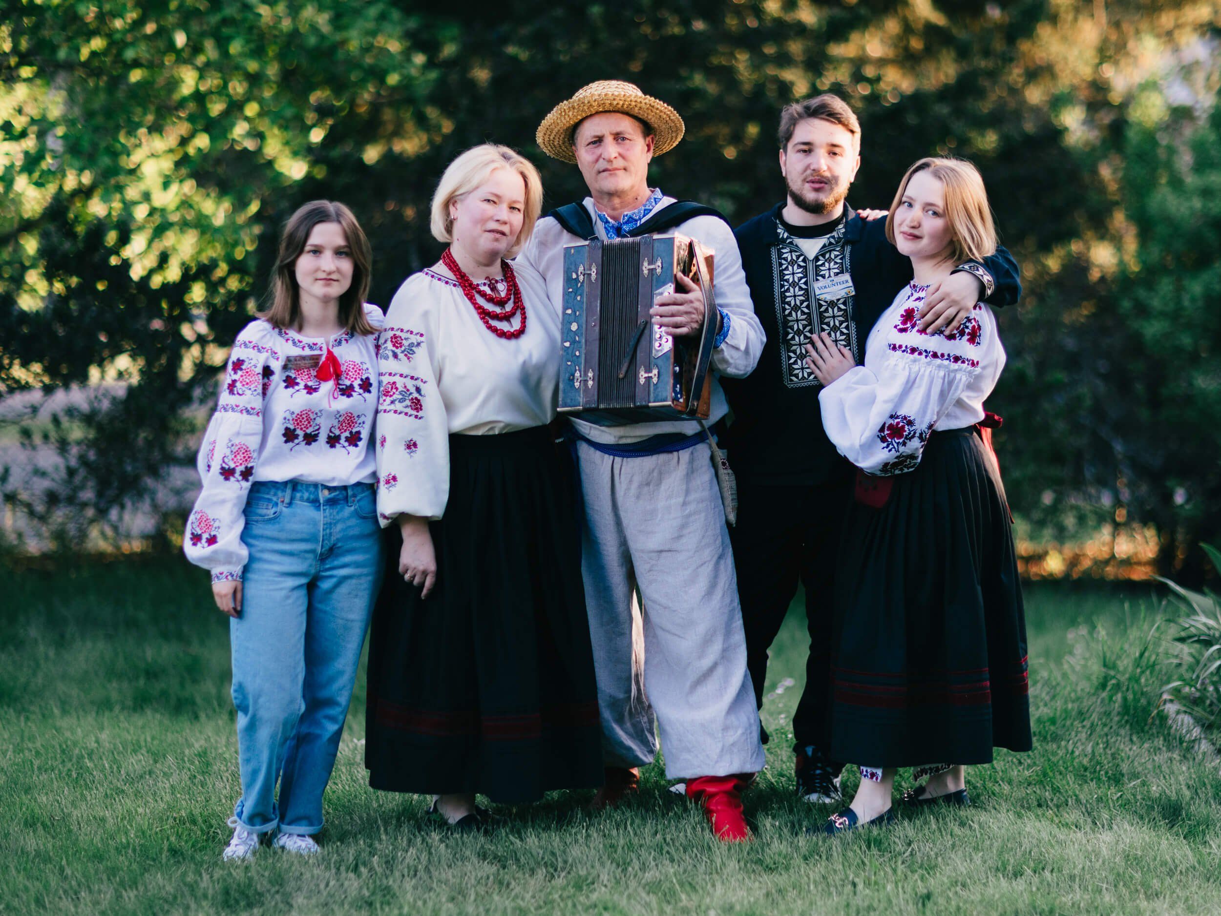 Five people in traditional Ukrainian embroidered clothing stand together outdoors at golden hour, with lush green foliage behind them