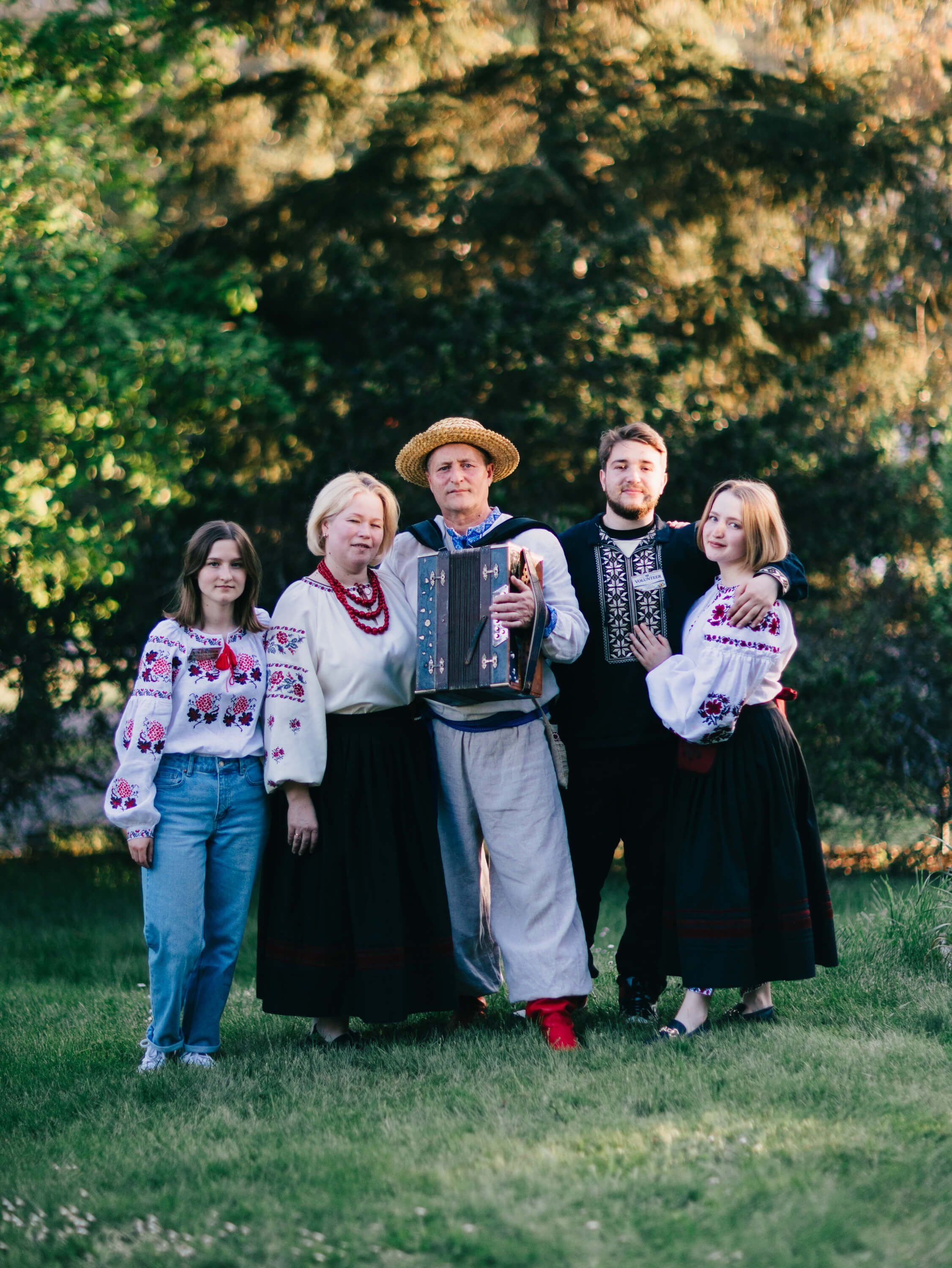 Five people in traditional Ukrainian embroidered clothing stand together on a grass lawn with ivy-covered stone wall glowing behind them in golden evening light