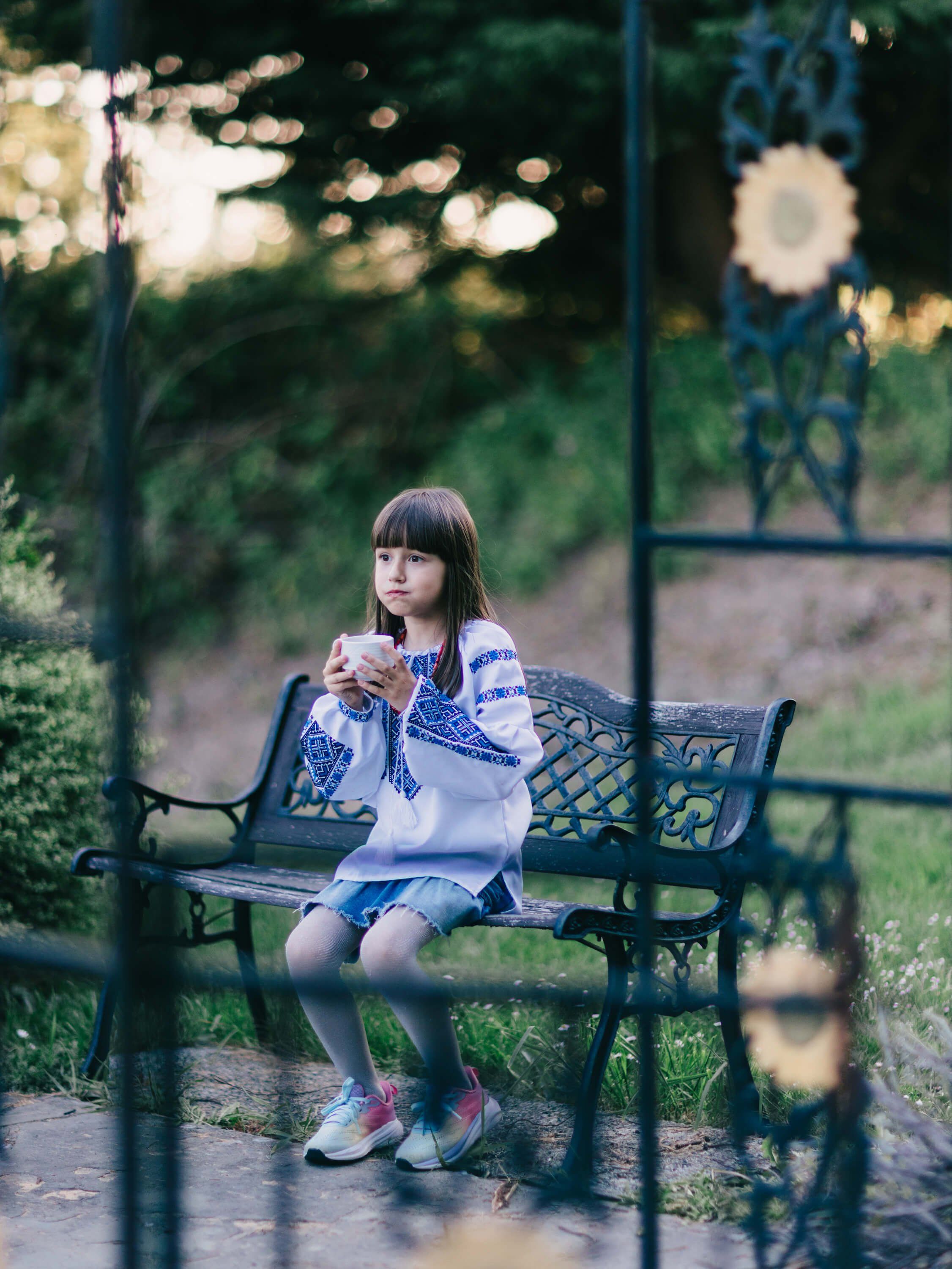 Young girl sitting on a bench behind a fence, holding a blue patterned fabric, golden hour light filtering through trees
