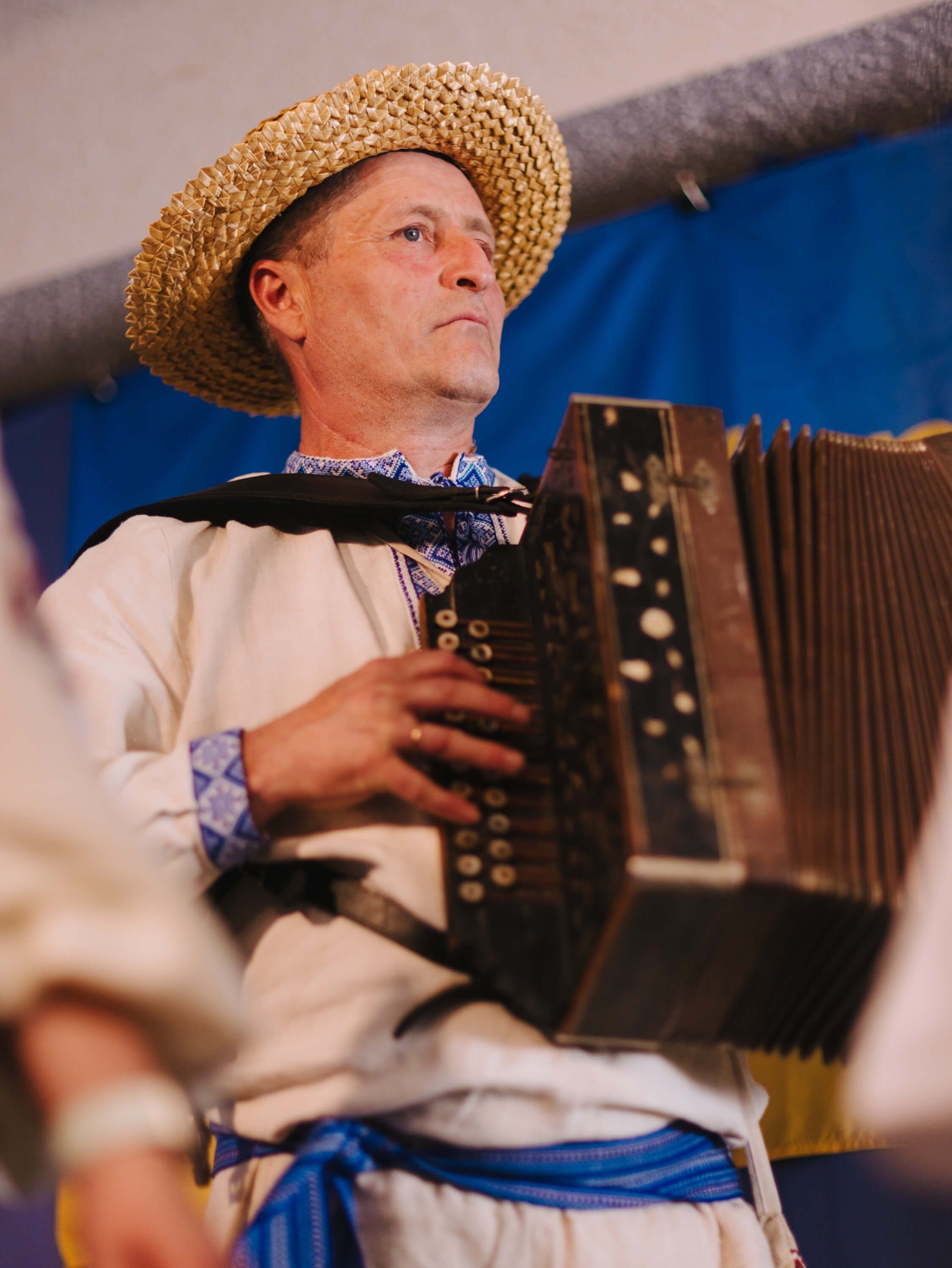 Musician in cream shirt and straw hat plays button accordion while gazing upward, blue fabric draped across his torso at an indoor fundraiser event