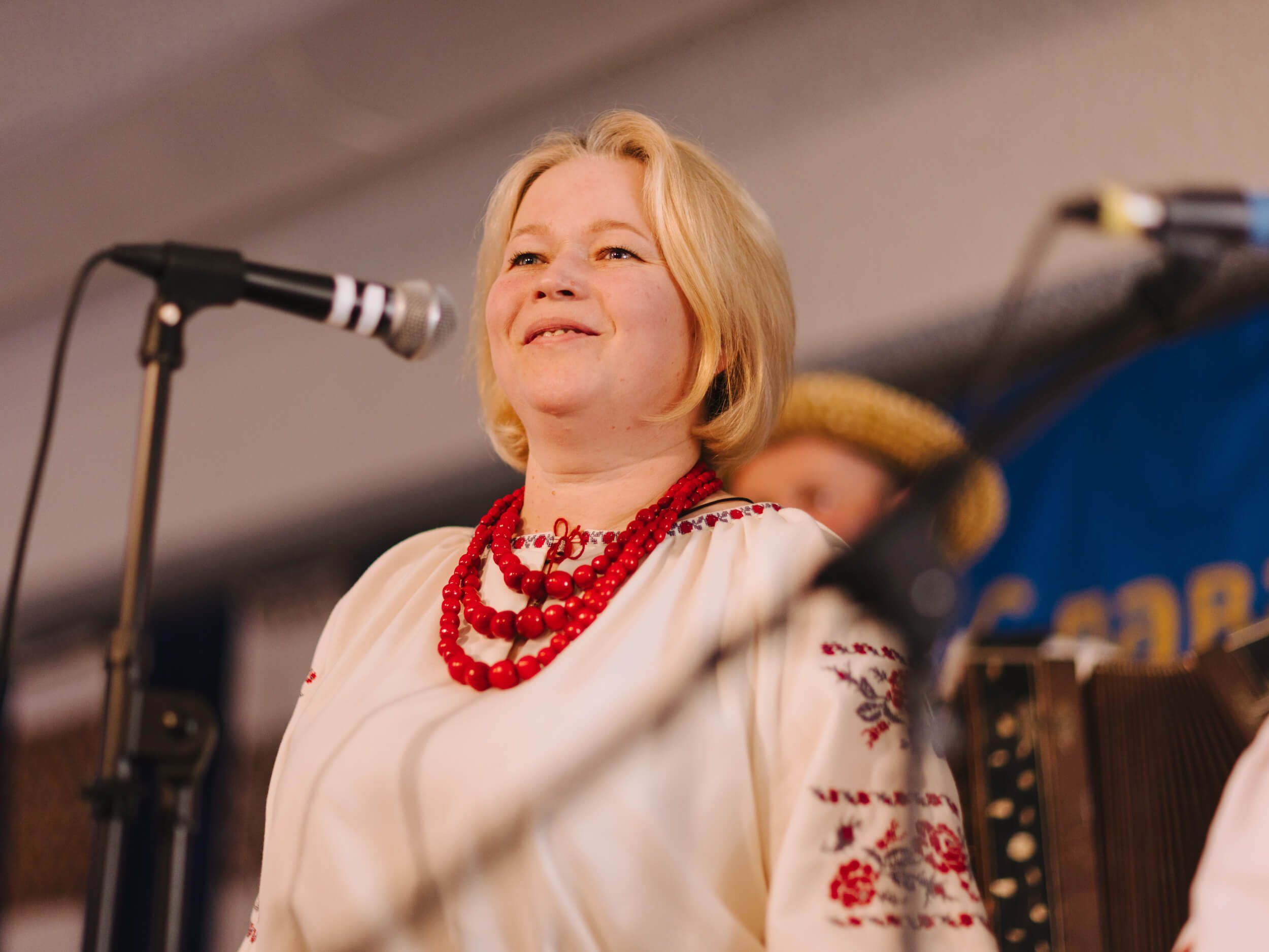 Woman in embroidered white linen and red beaded necklace singing at microphone during Ukrainian fundraiser dinner