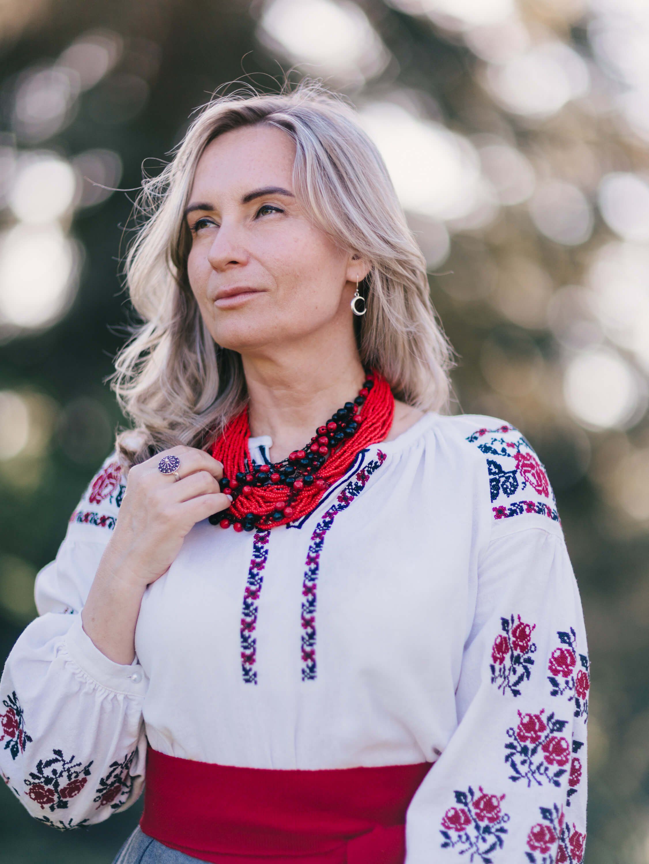 Woman in embroidered Ukrainian blouse and red scarf gazes thoughtfully into distance, soft natural light filtering through trees behind her