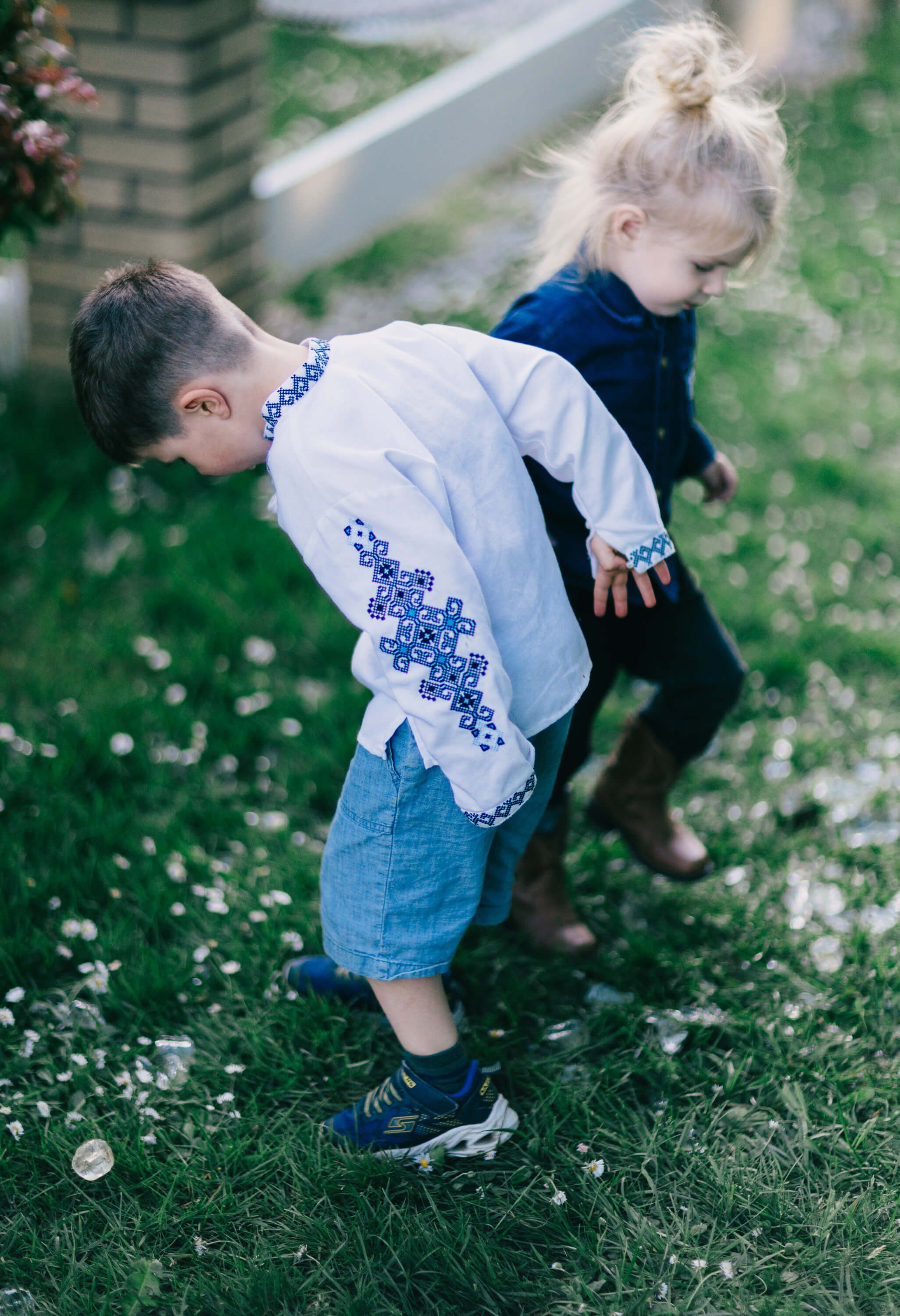 Two young children play on grass, one in a white embroidered shirt bending forward, the other in dark clothing behind, captured in warm afternoon light