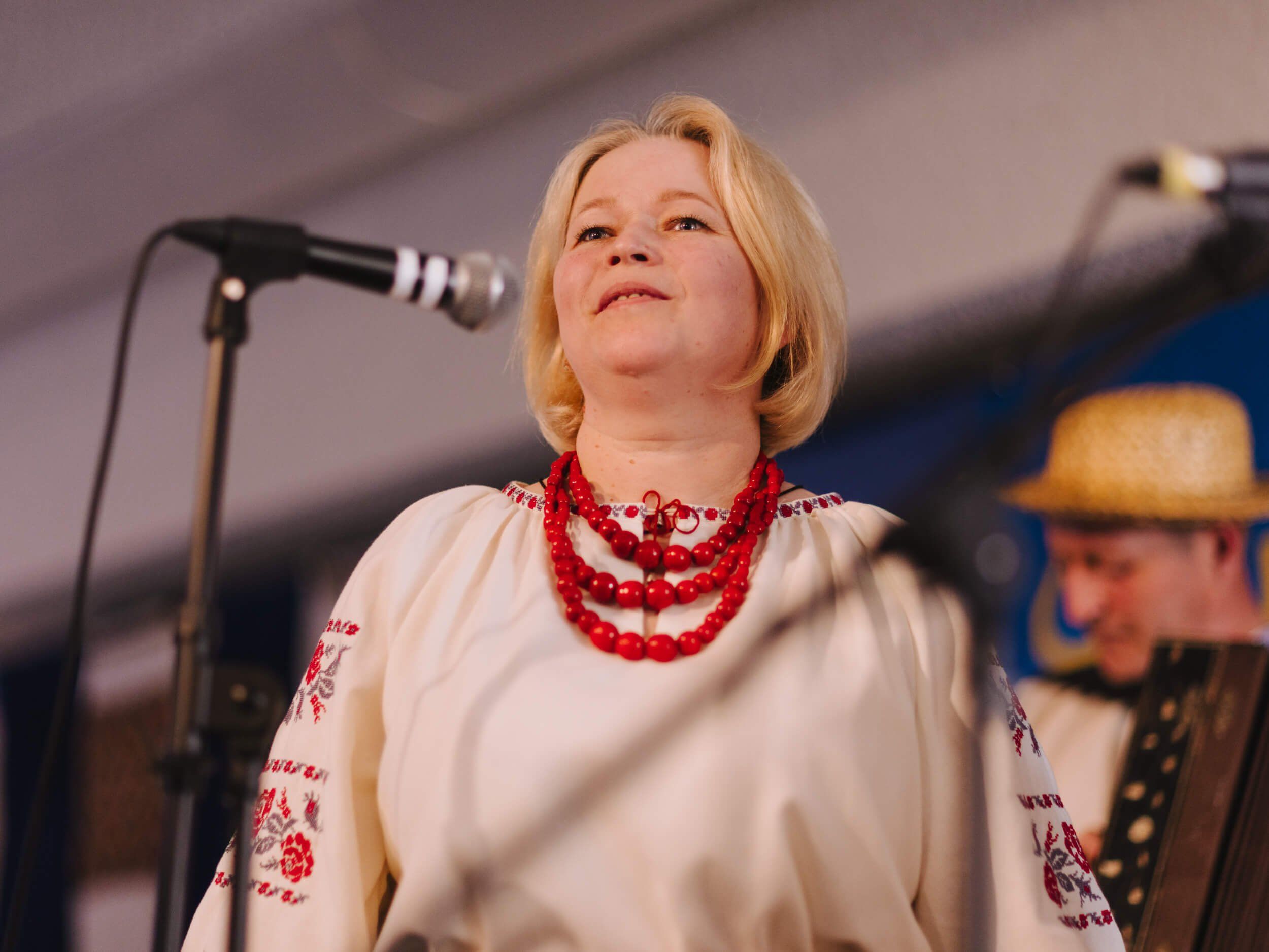 Woman in embroidered white blouse and red beaded necklace singing at microphone during Ukrainian fundraiser dinner
