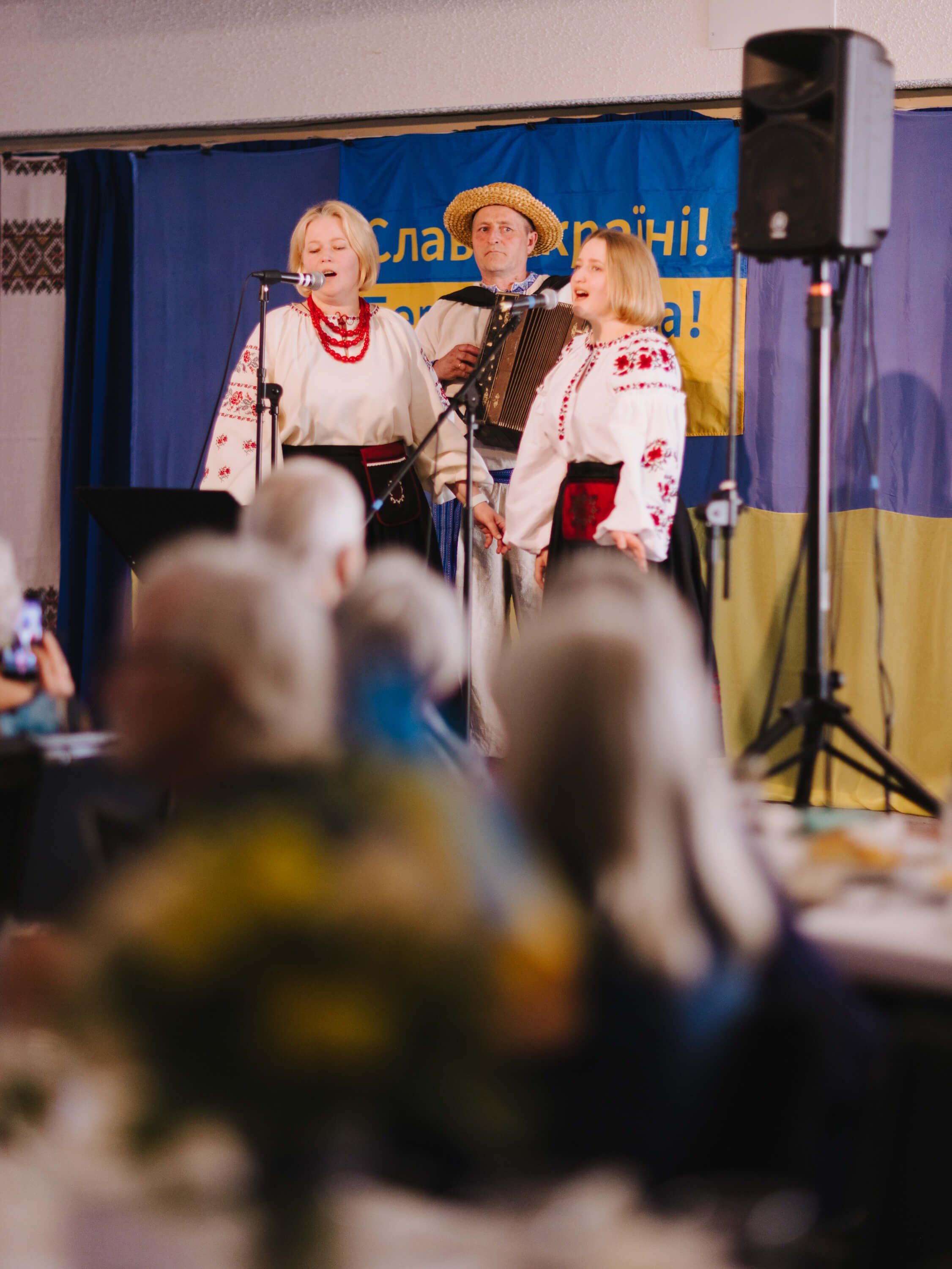 Three women in embroidered Ukrainian traditional dress perform at a microphone on stage during an indoor fundraiser dinner