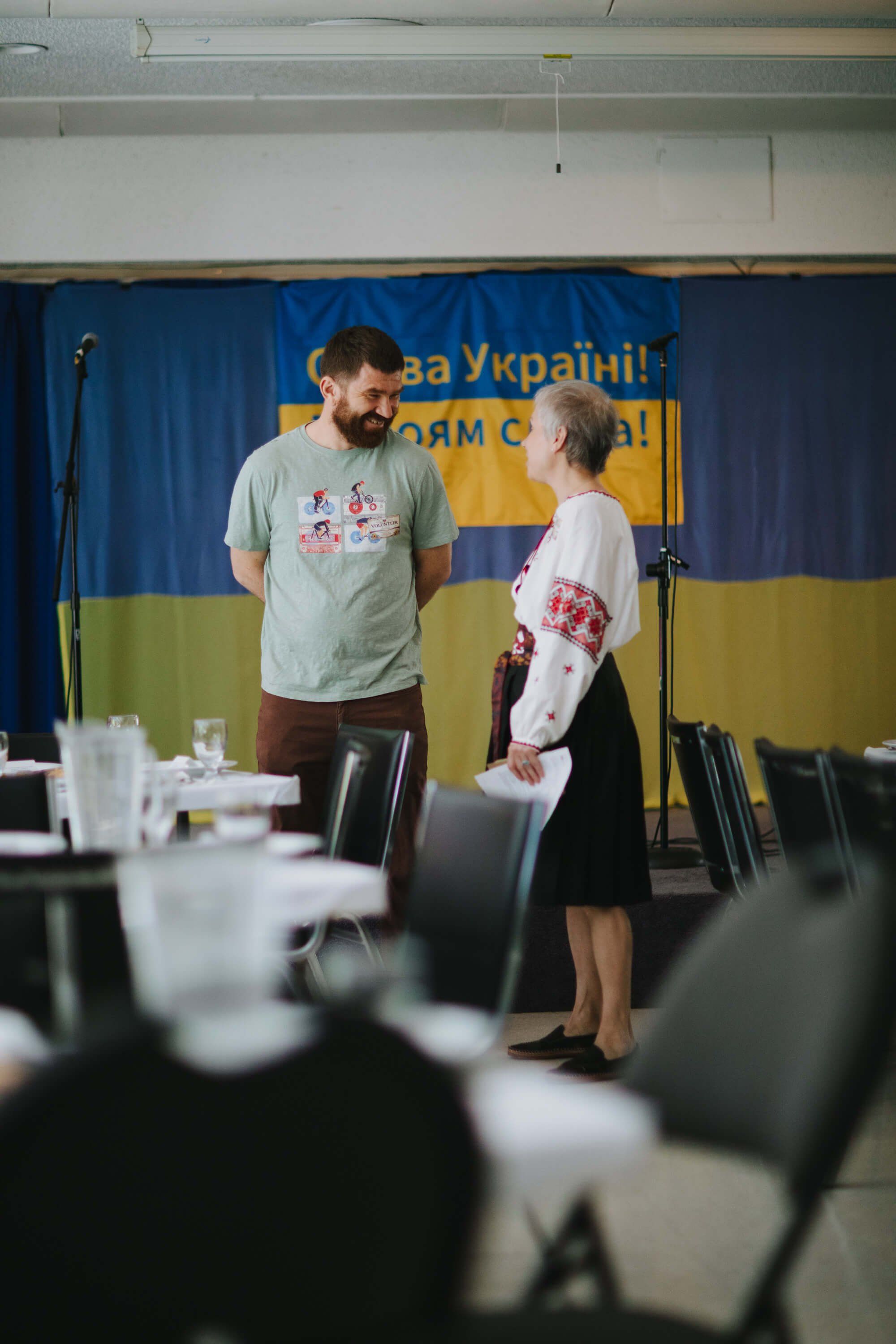 Two people in embroidered white shirts speak on stage before a blue and yellow Ukrainian flag backdrop at an indoor fundraiser dinner