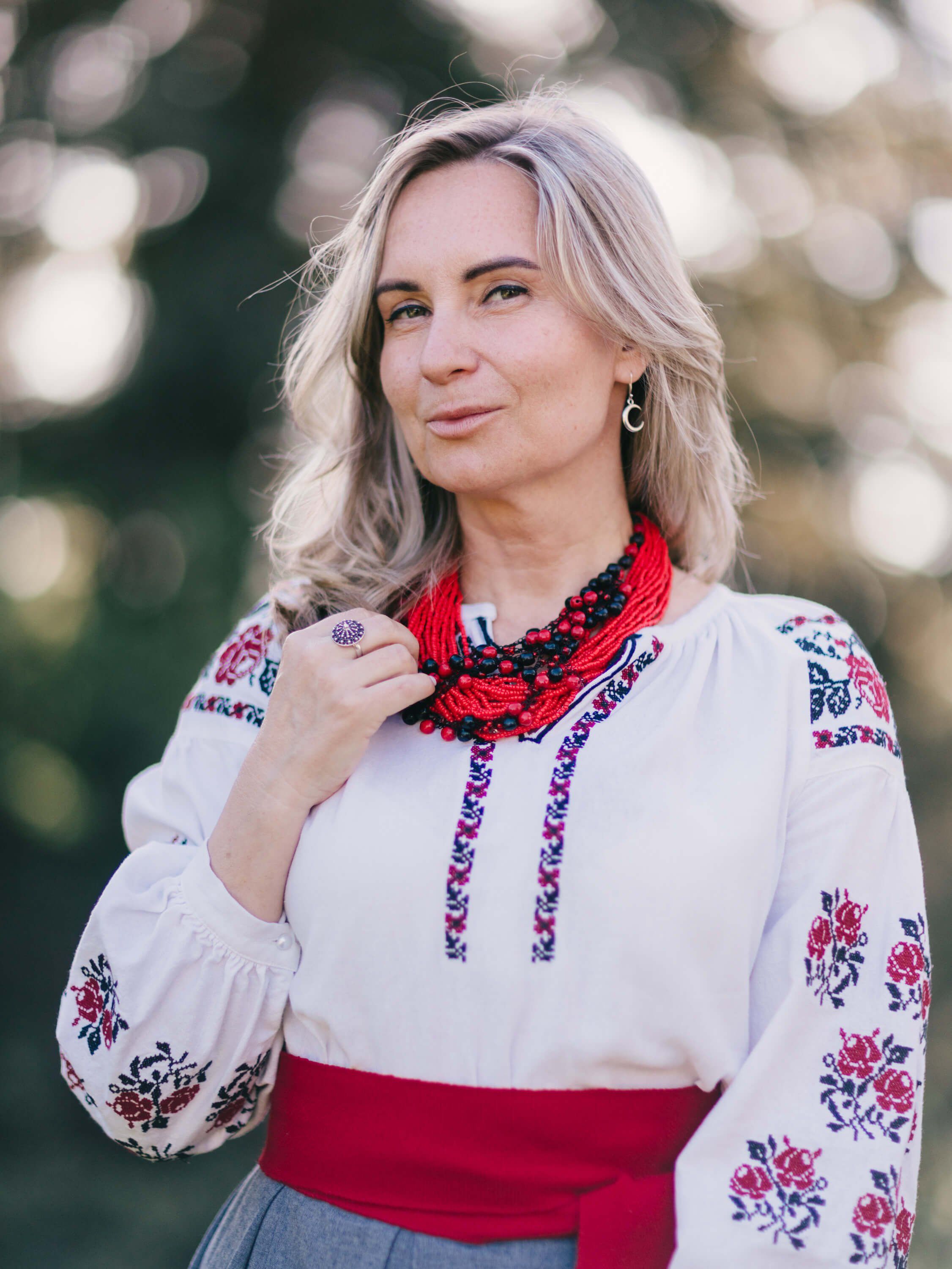Woman in embroidered Ukrainian dress with red beaded necklace smiles at camera with soft garden bokeh behind her