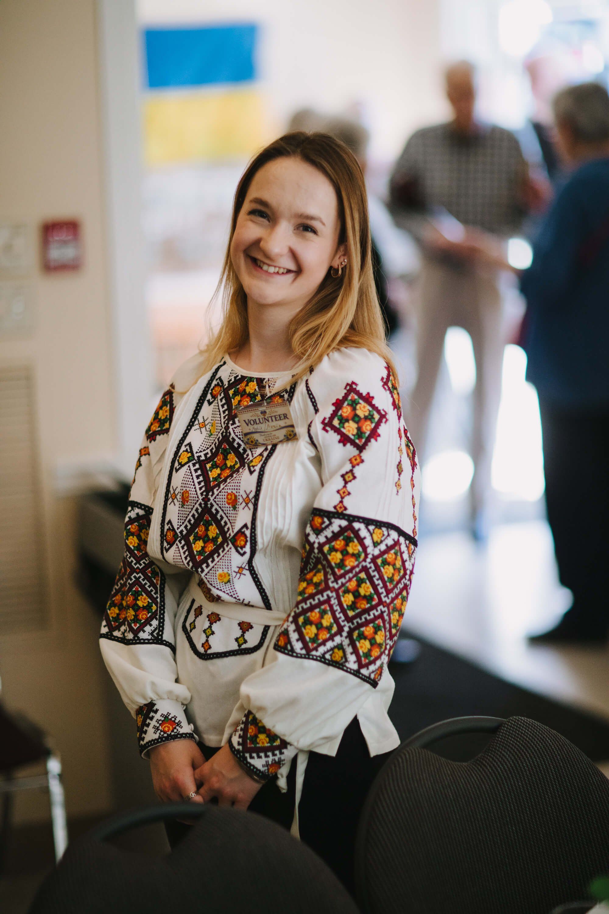 Smiling woman in embroidered Ukrainian blouse at indoor event with blurred guests and Ukrainian flag visible behind her