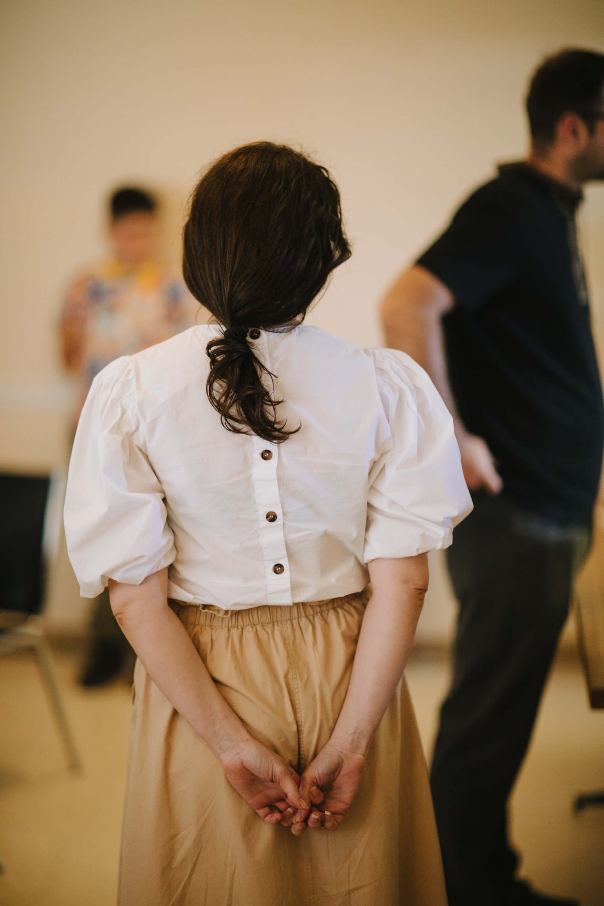 Woman in cream blouse and tan skirt stands with hands clasped, back to camera, at indoor fundraiser event