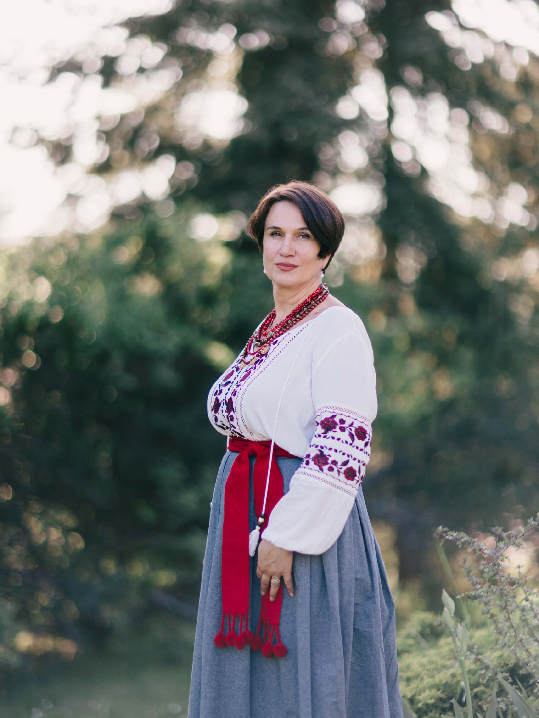 Woman in traditional Ukrainian embroidered white blouse and grey skirt with red sash stands among evergreen trees in golden evening light